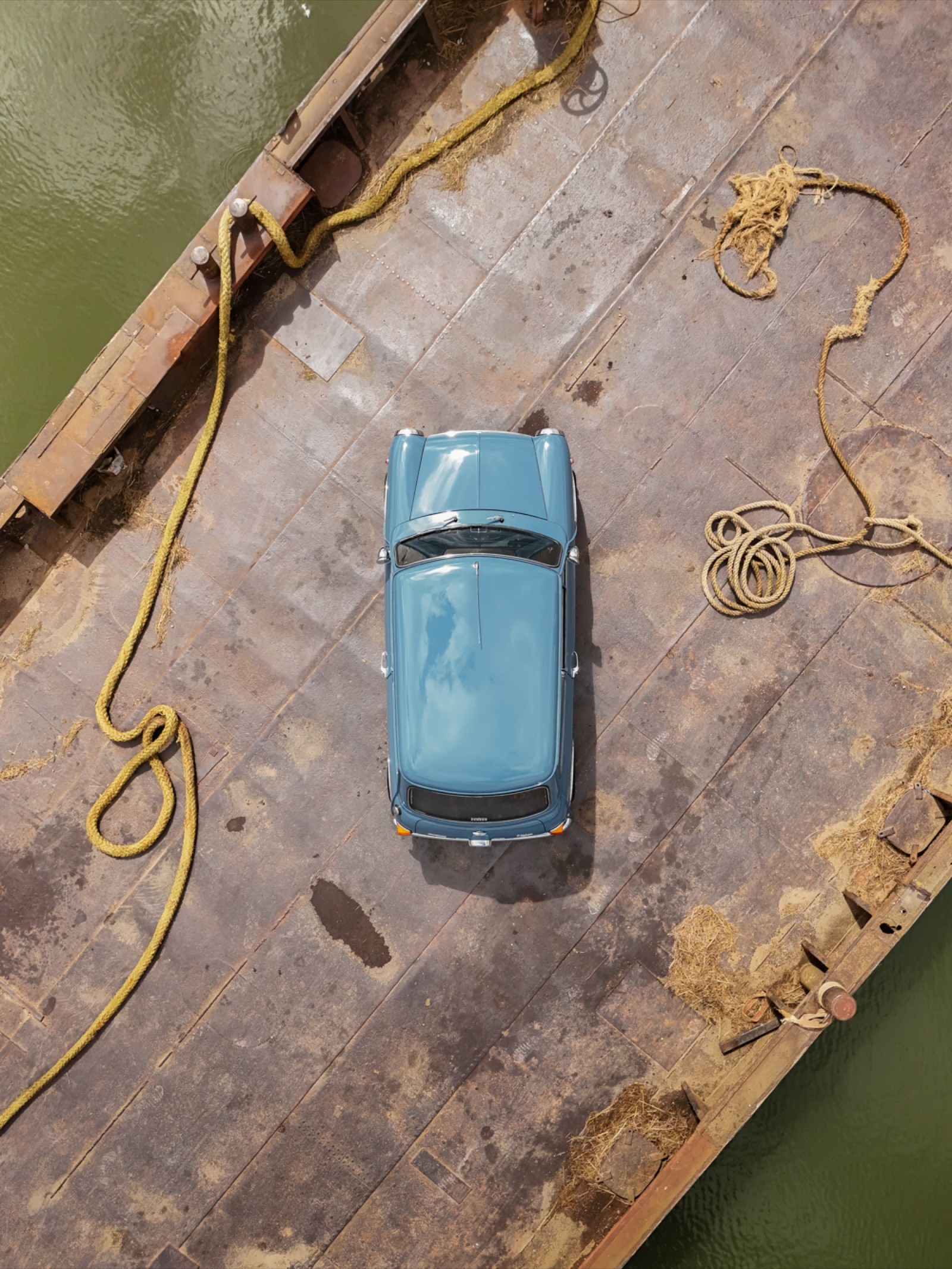 A small blue toy car resting on a wooden surface beside calm water.