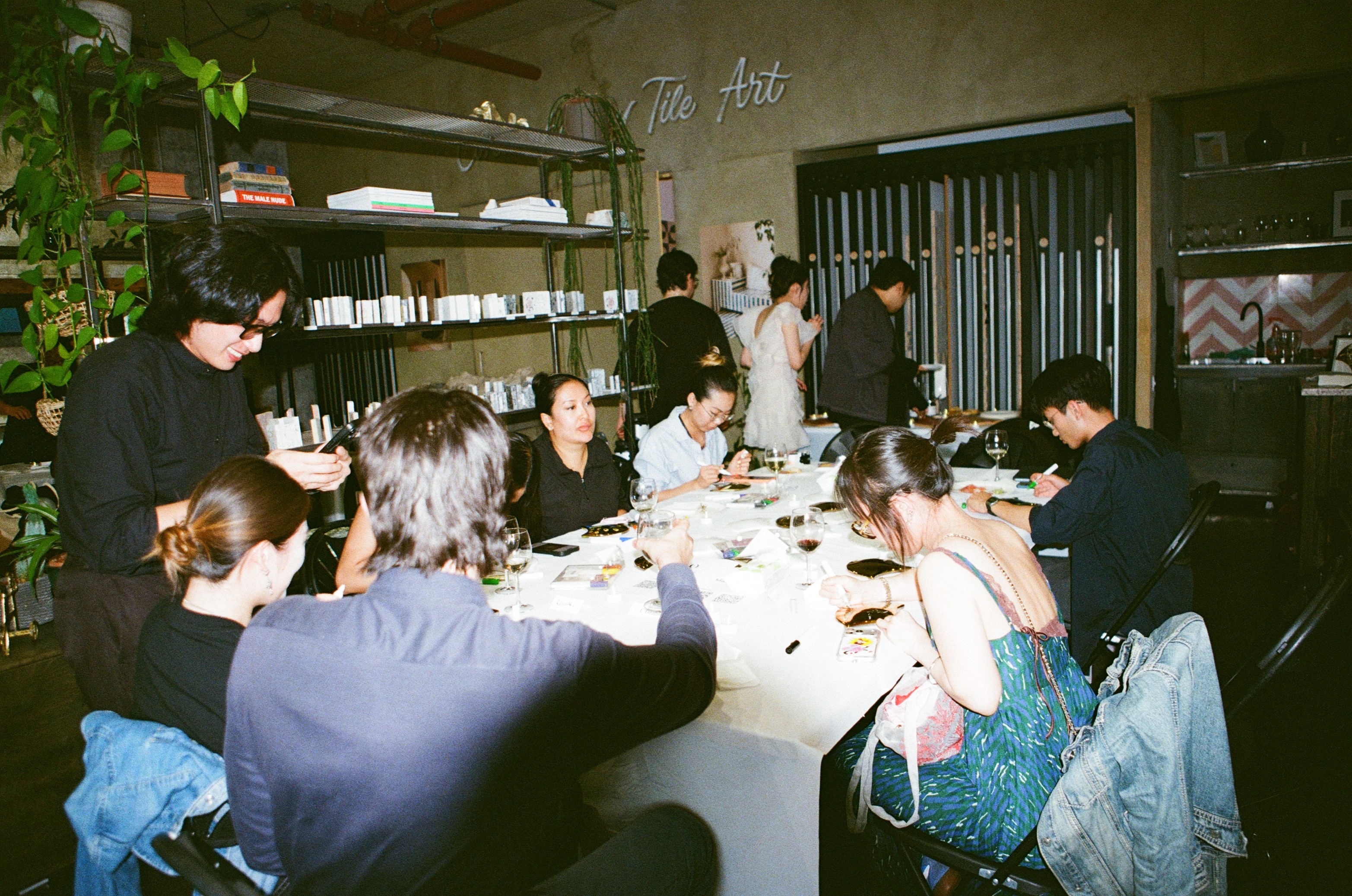Guests seated along a long white-clothed table in the Otto Tiles showroom, painting tiles with markers, wine glasses and paint trays between them, a founder standing at the left with a phone, planting and stacked sample shelves behind, a neon sign reading Tile Art on the far wall.