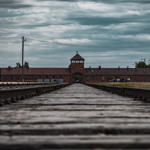 Houten spoorrails die leiden naar een bakstenen gebouw met een centrale ingangstoren onder een bewolkte hemel.
