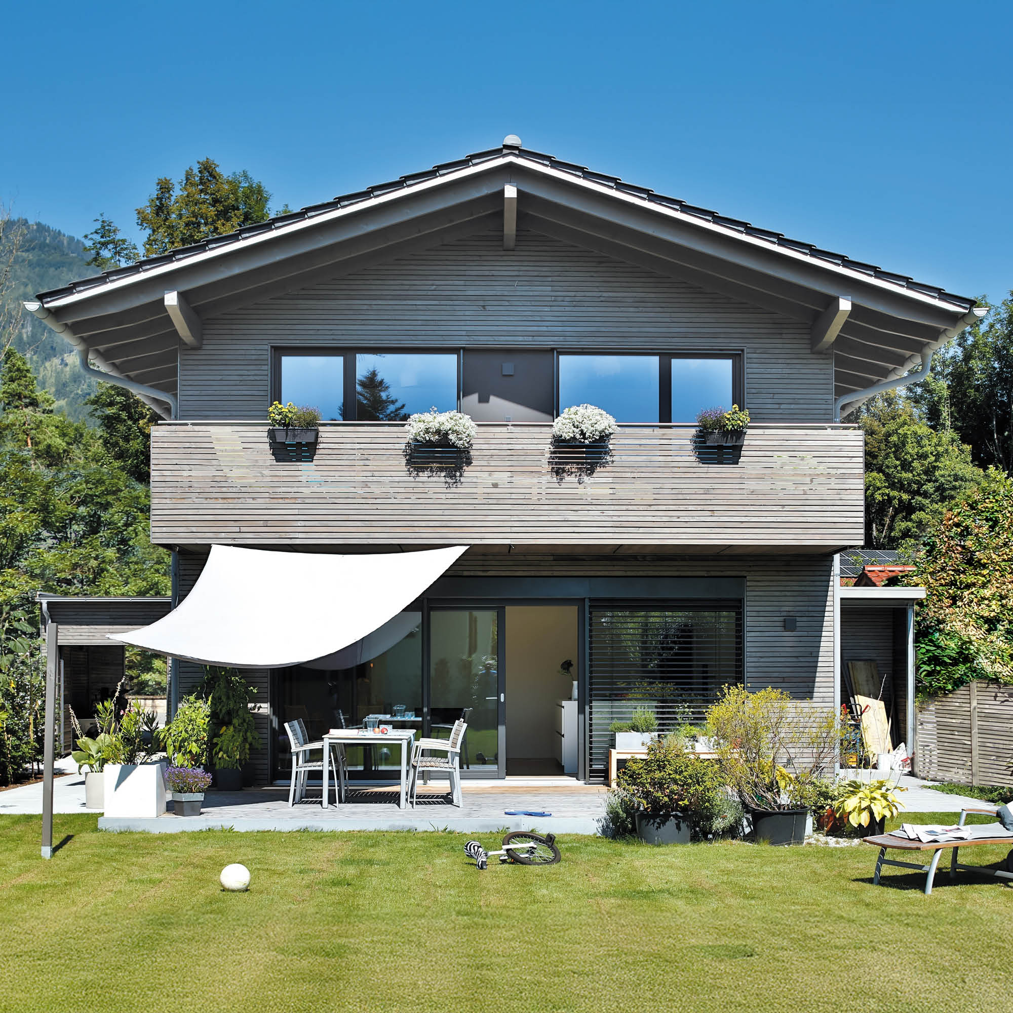 Modernes Holzhaus mit Balkon, Terrasse, Sonnensegel und parkähnlichem Garten.