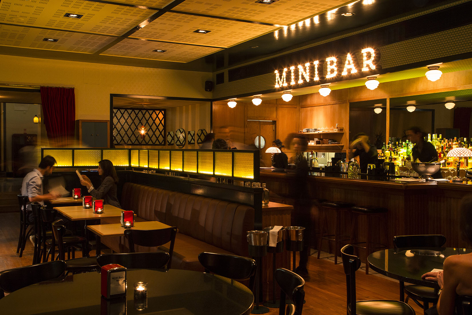 A dimly lit bar with a long wooden counter, stools, and "MINIBAR" in neon letters on a dark wall above the service area.