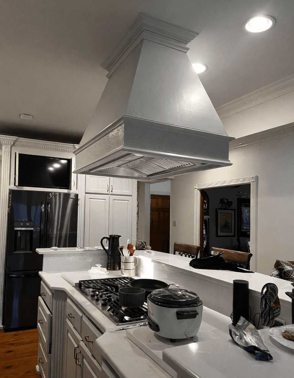 A modern, glass-walled house with warm interior lighting is perched on a rocky cliff overlooking a calm sea at sunset, under a clear blue sky.