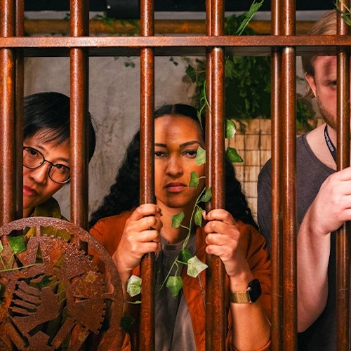 Drie personen staan achter roestige metalen tralies, met groene ranken die om de tralies verstrengeld zijn.