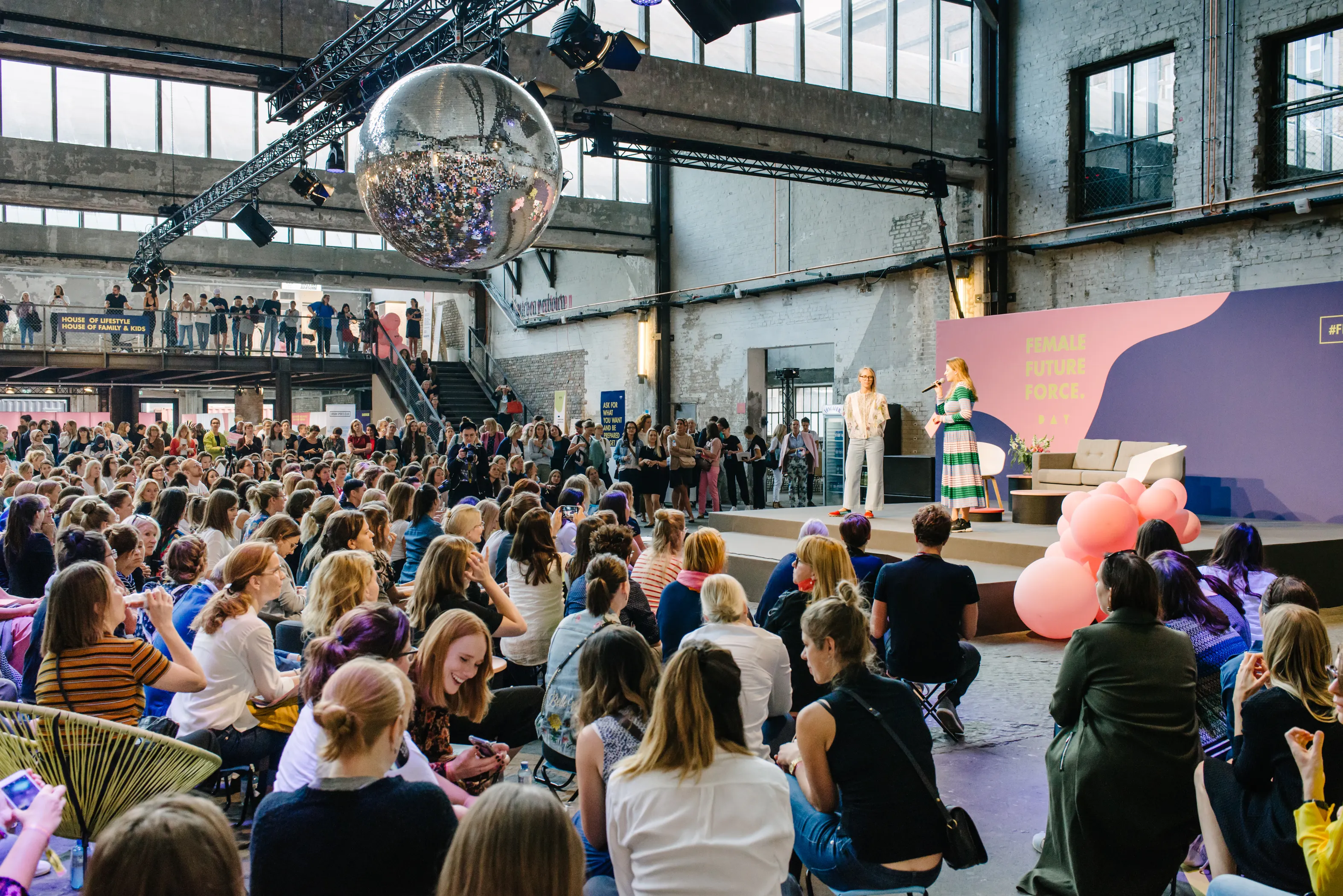 Crowded hall at the FEMALE FUTURE FORCE DAY with a speaker on stage, purple backdrop wall, pink sculpture and a large disco ball overhead.
