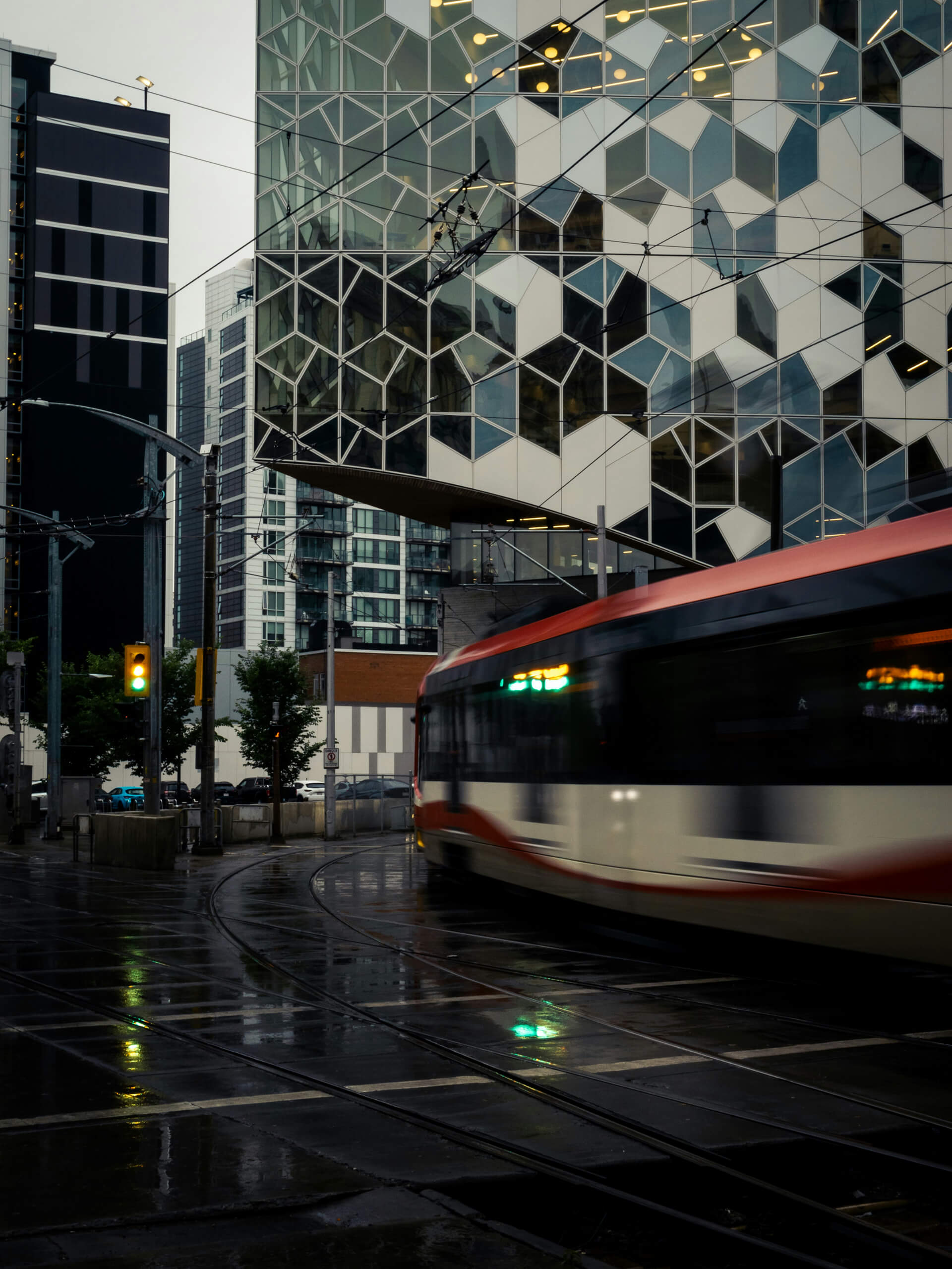 A red and white light rail train blurs through a rainy urban intersection in front of Calgary’s Central Library, known for its striking hexagonal glass façade. Reflections from traffic lights and surrounding buildings shimmer on the wet pavement, adding depth to the moody cityscape.