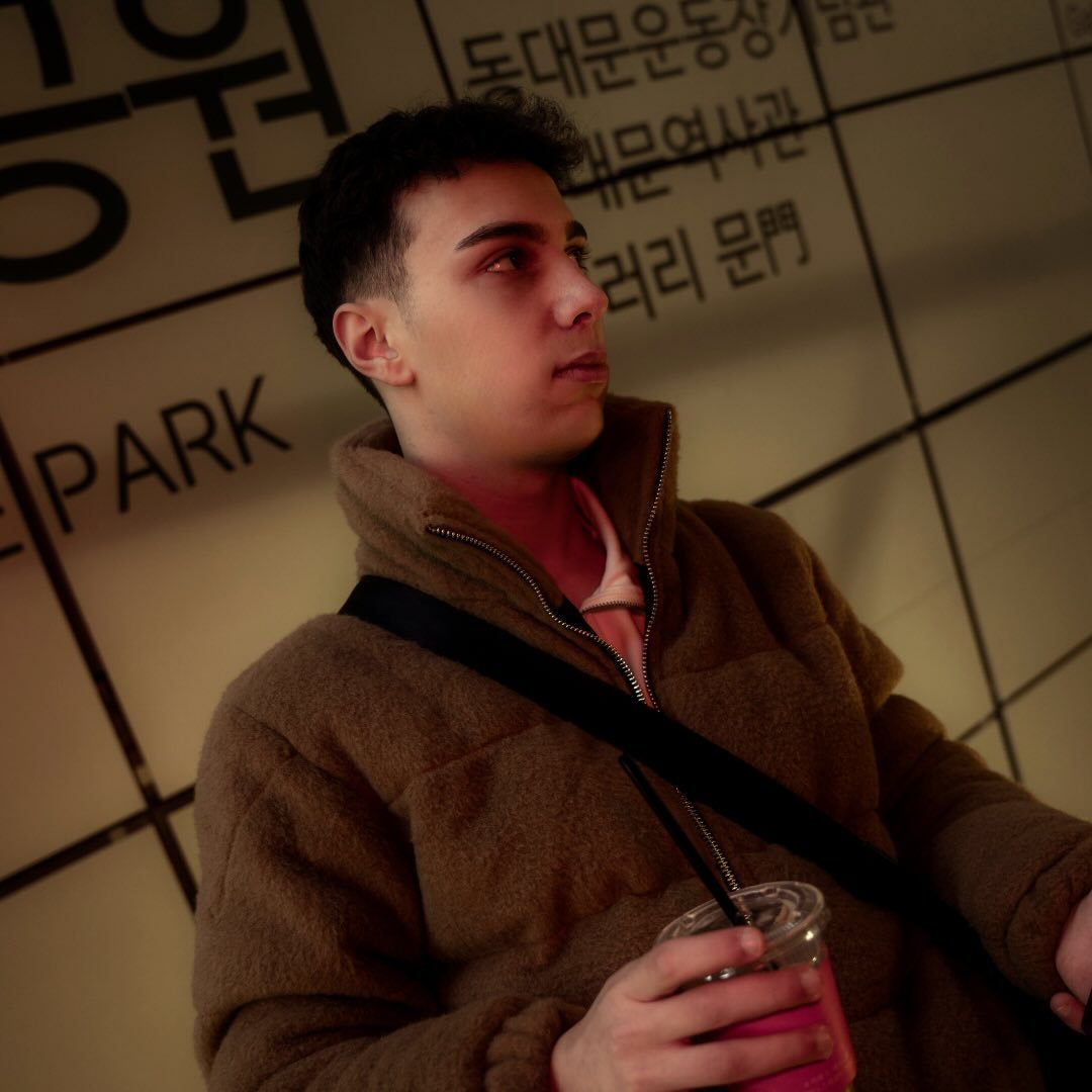 A smiling person with curly hair sits at a desk, looking at a laptop against a dark background.