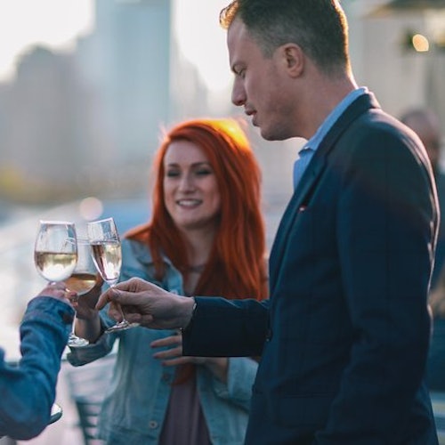 Three people outdoors toasting with glasses of white wine. A man in a suit and a woman with red hair are in focus.
