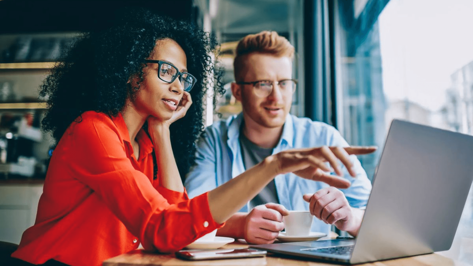 Two applicants sitting together at a café table, discussing and pointing at a laptop screen