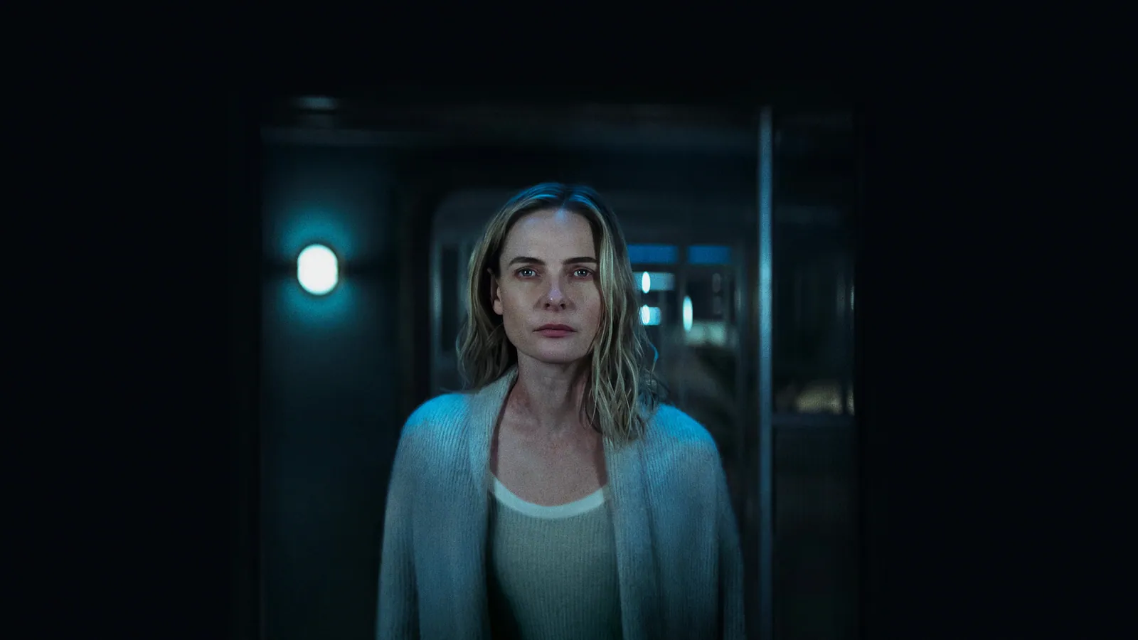 A woman with blonde hair stands in dim lighting, wearing a light-coloured sweater, creating a mysterious and contemplative atmosphere.