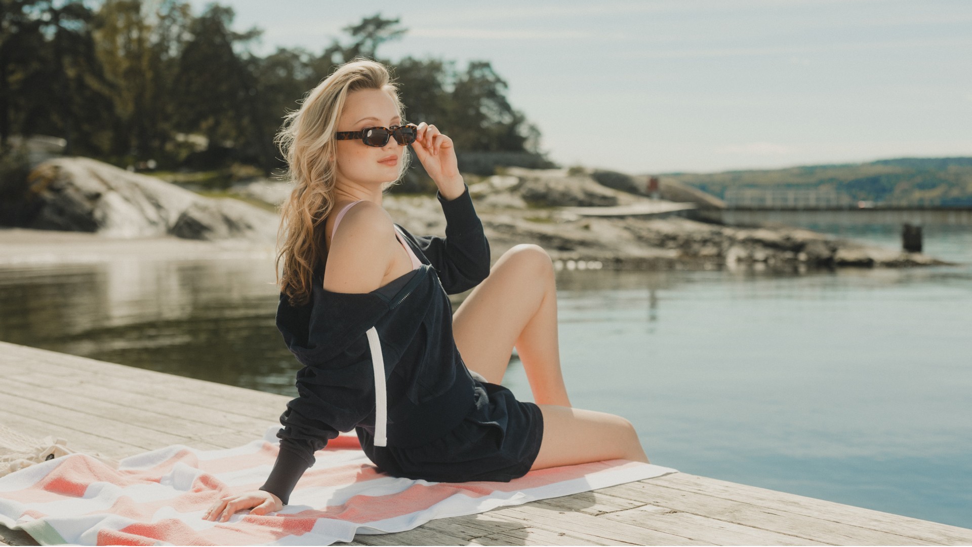 Young woman sitting outside by the ocean, wearing a navy ziphood, navy shorts and dark sunglasses