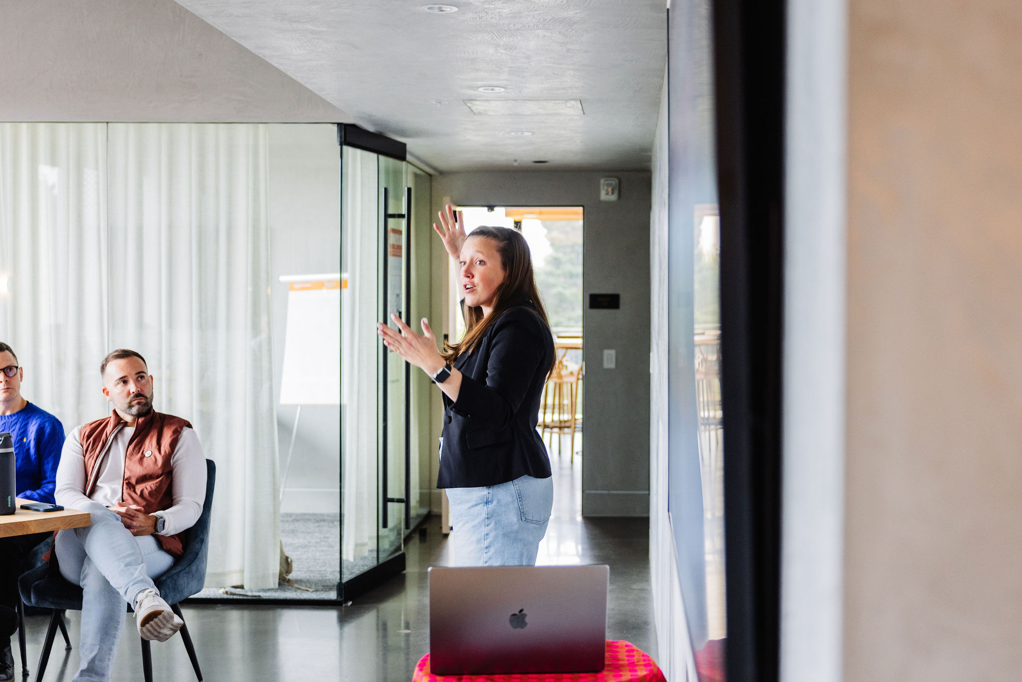 Woman speaking and gesturing during a presentation in a modern office space, with coworkers seated nearby.