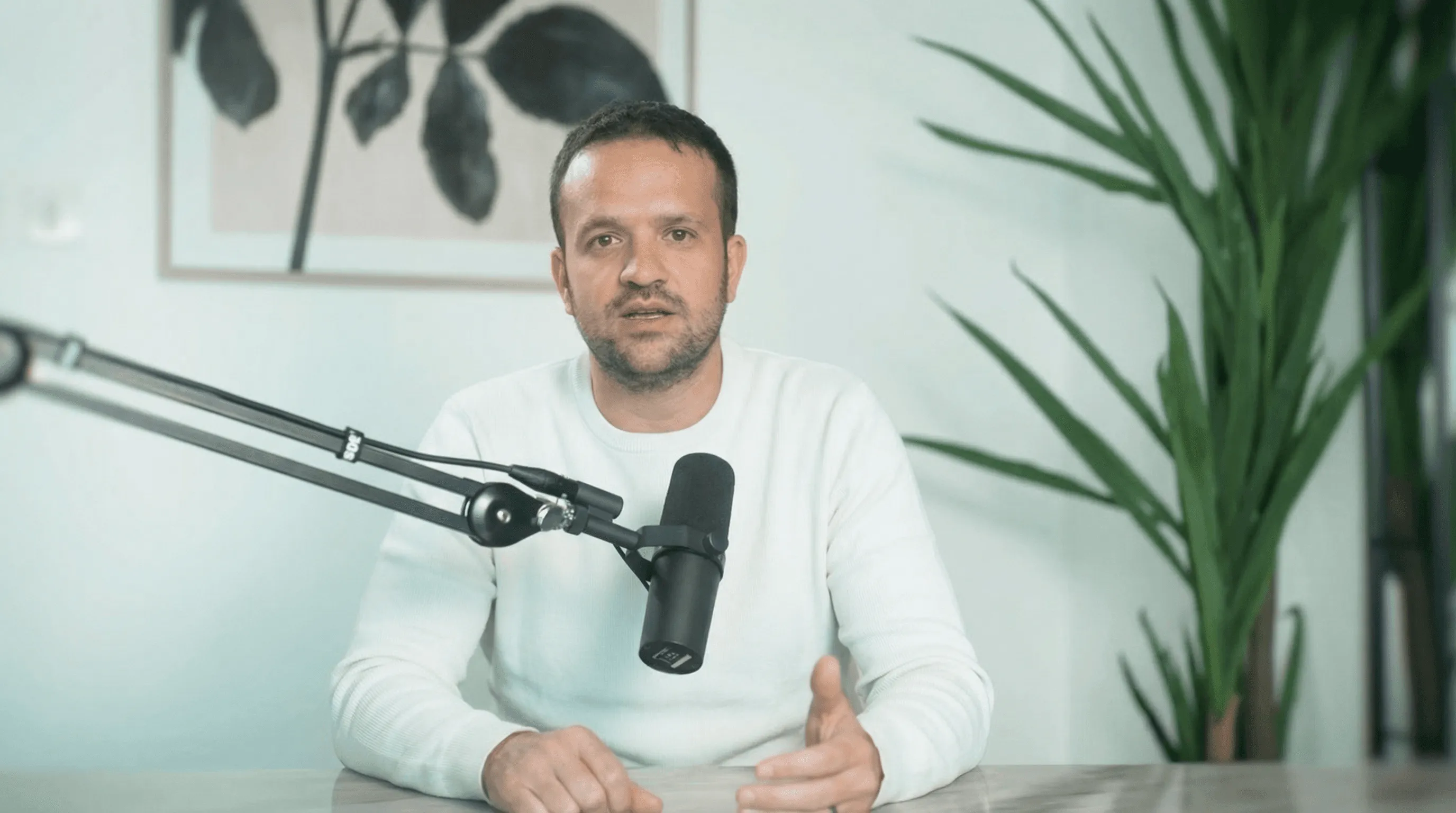 Persona hablando a cámara frente a un micrófono de estudio, sentada en una mesa, en un espacio interior minimalista con plantas y arte en la pared.