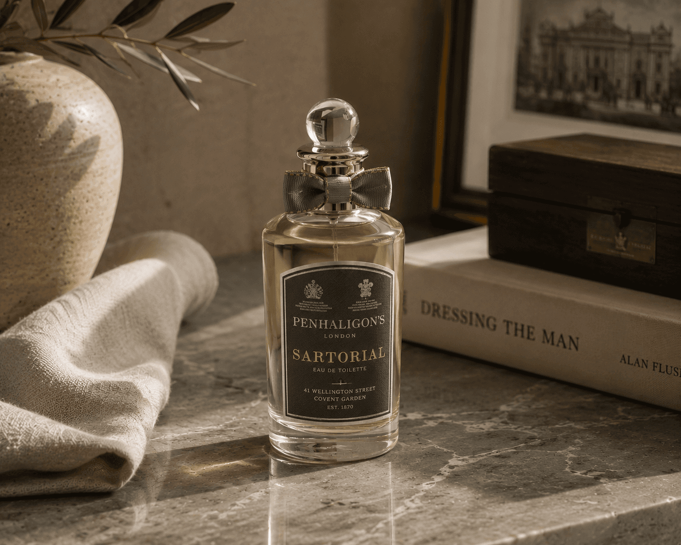 A bottle of Penhaligon's Sartorial Eau de Toilette featuring a clear round glass body, dark gray label with yellow lettering, a circular glass stopper, and a gray fabric bow tied around the neck. The perfume rests on a polished gray marble surface next to a book titled "Dressing the Man", a dark wood box, a ceramic vase with olive branches, and folded linen cloth.