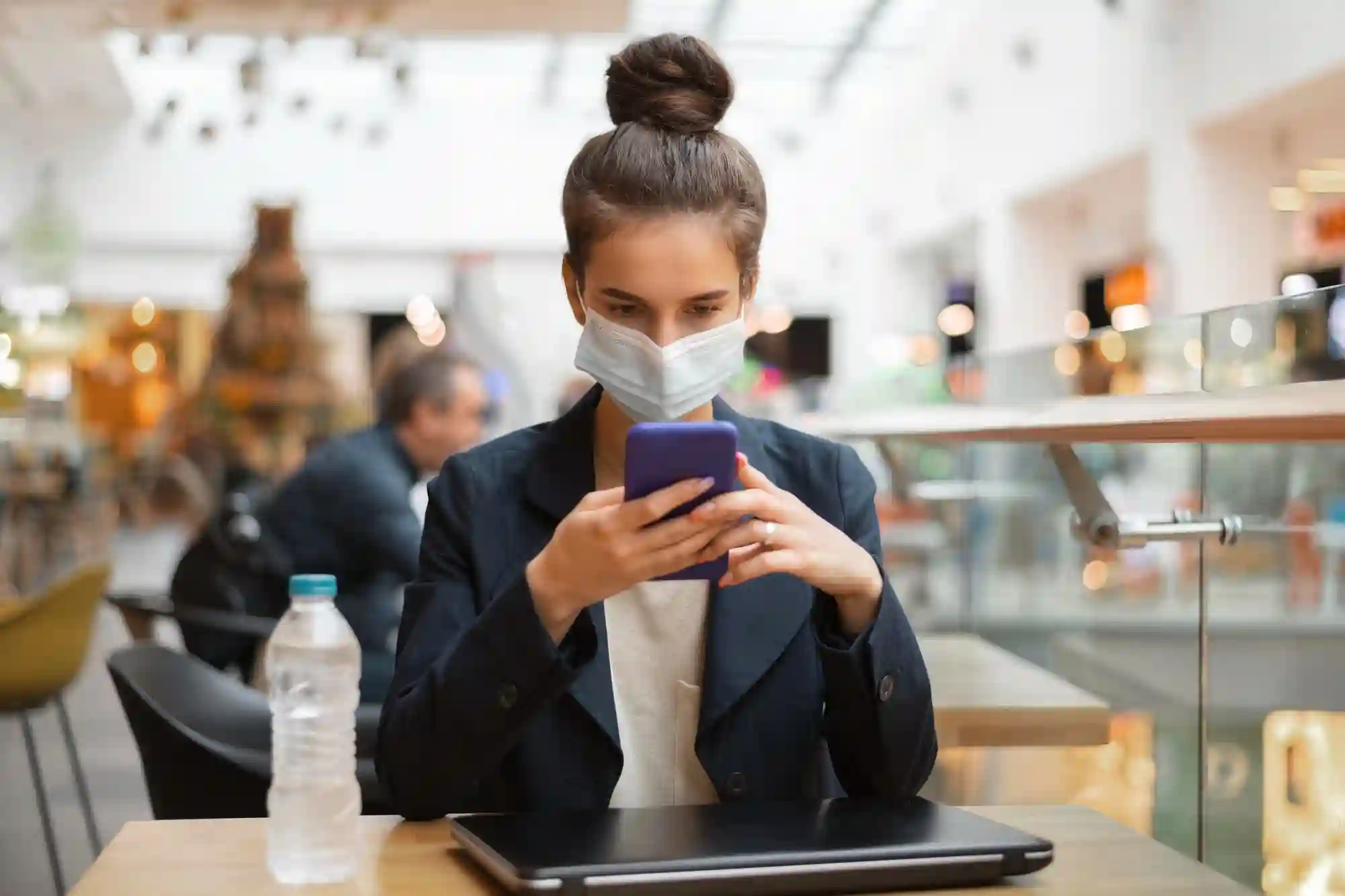 A woman wearing a face mask uses her smartphone in a public space, representing digital communication during the pandemic.