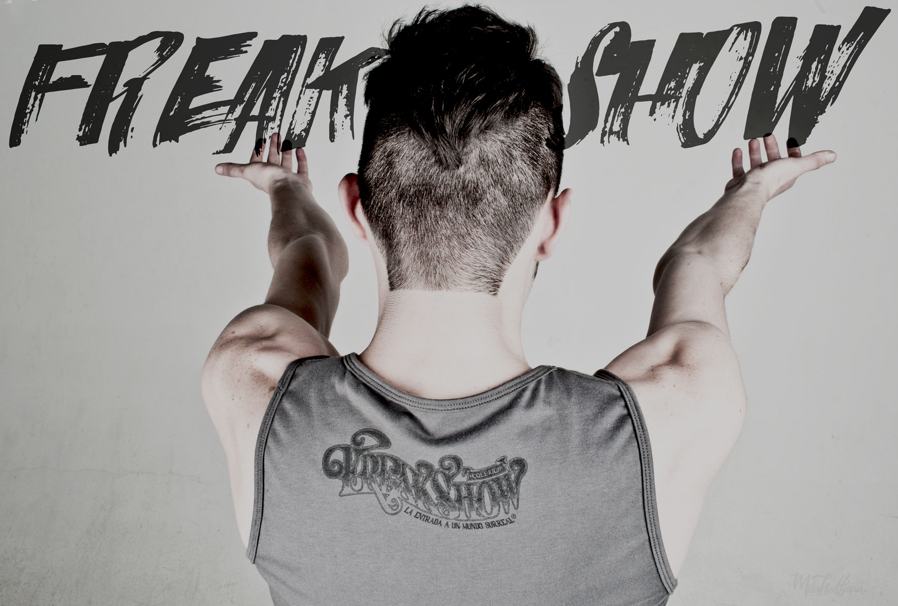 Back view of a man in a grey tank top with his arms raised, showing a "FREAK SHOW" logo on the garment. Large "FREAK SHOW" text is integrated into the grey background.