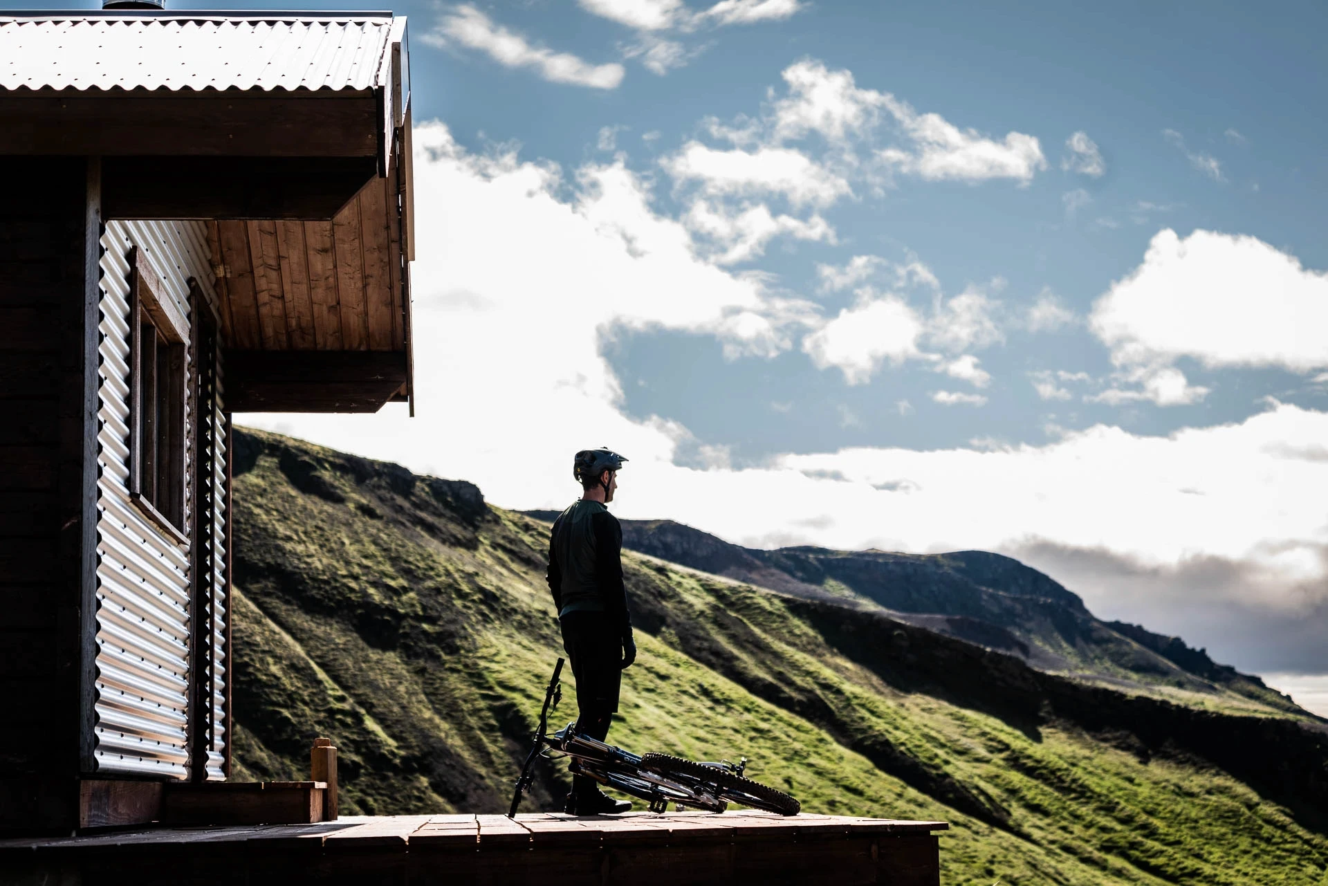 Cyclist standing on a cabin porch beside a bike, overlooking green mountains.