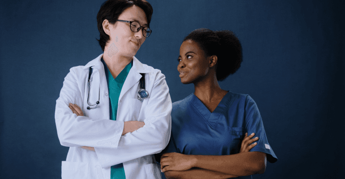 A doctor in a white lab coat and stethoscope stands with arms crossed alongside a nurse in blue scrubs, both smiling at each other against a dark blue background.