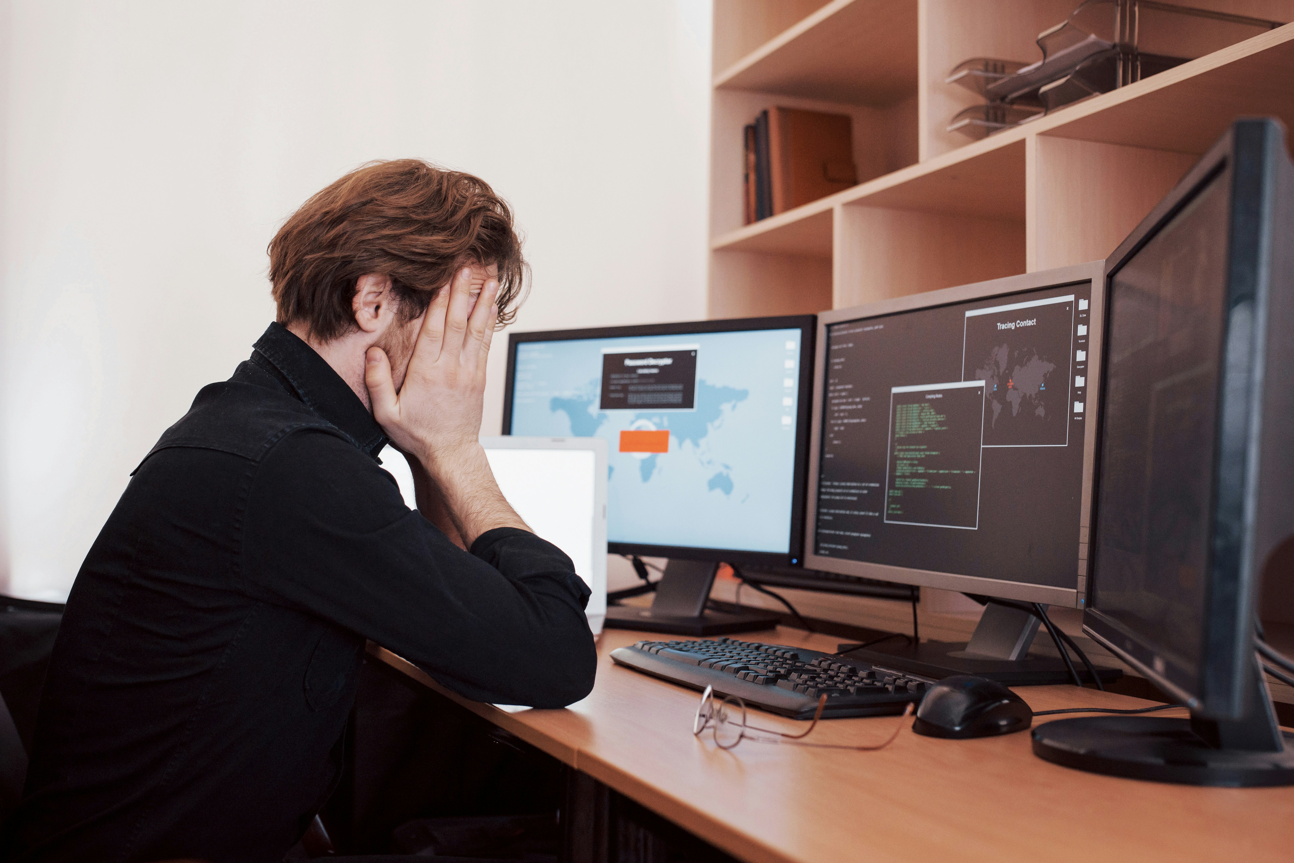 A professional sitting at a desk with head in hands, facing multiple computer screens, representing stress and uncertainty in modern work environments.