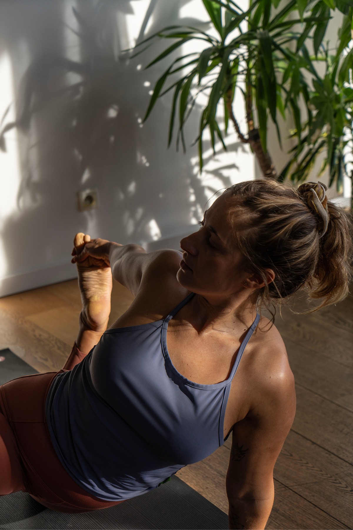 A woman practices yoga on a wooden floor in a sunlit room, with lush green plants adorning the background.