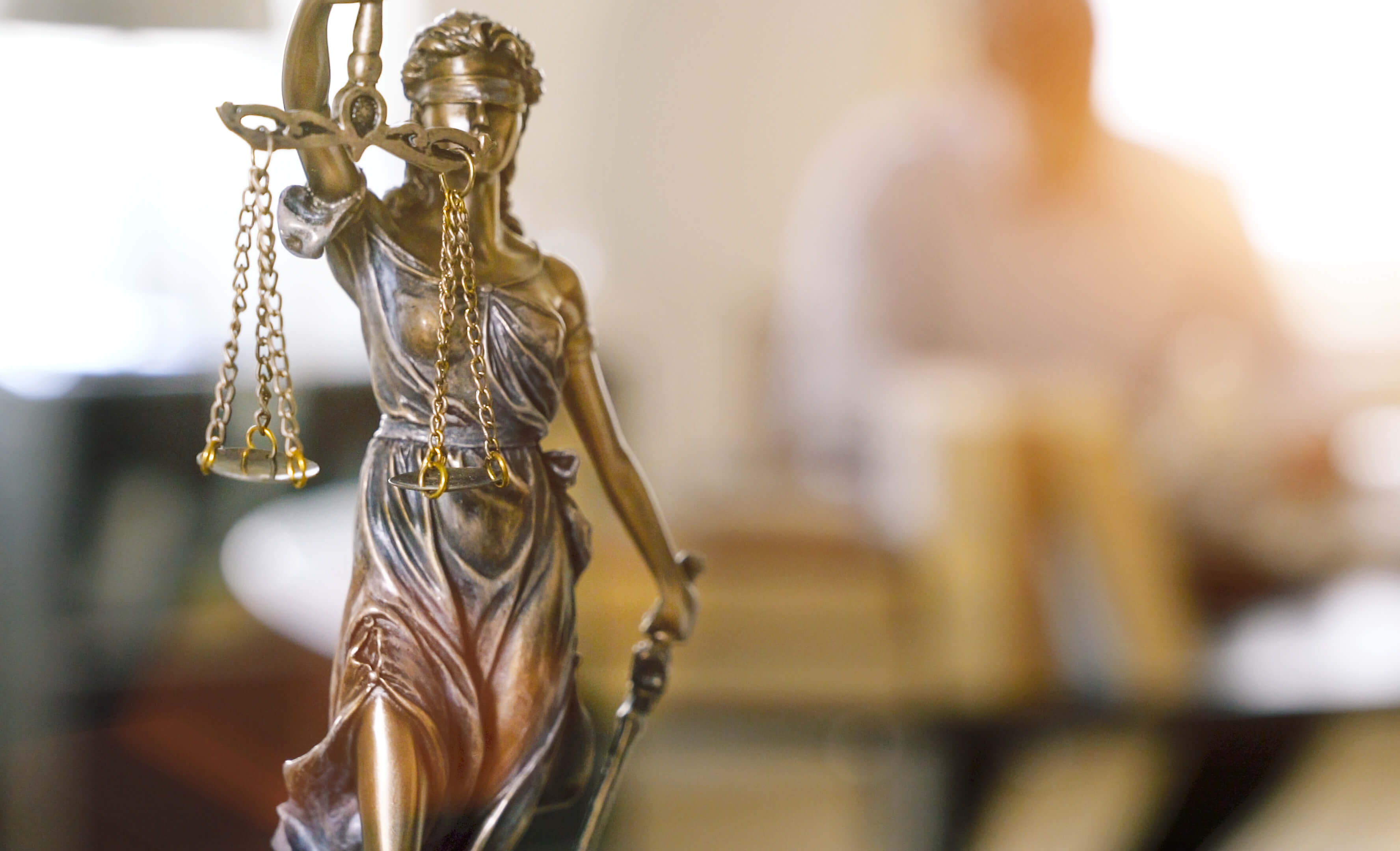 Um close-up de uma estátua da Dama da Justiça em primeiro plano, com uma pessoa desfocada e o fundo de um escritório ao fundo.
