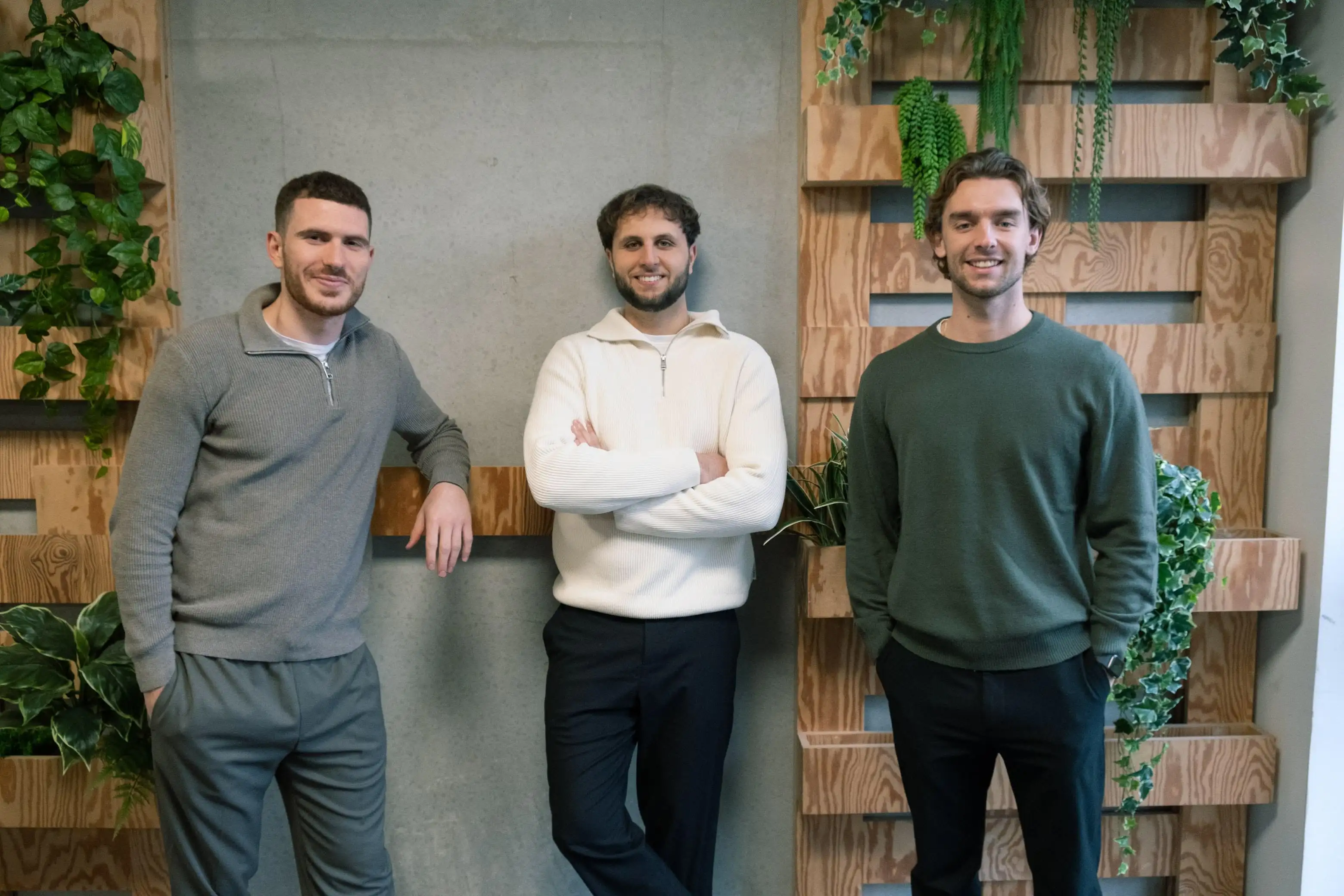 Three men are standing together, smiling, in a cozy indoor space with greenery and a wooden backdrop.
