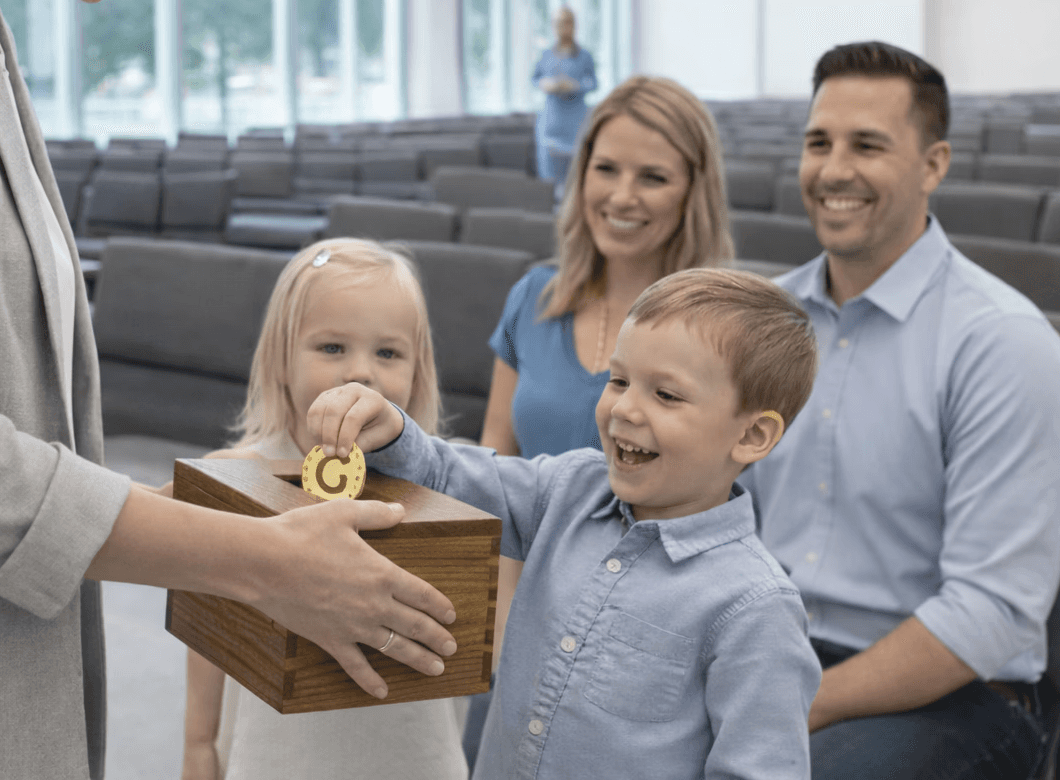 Una persona con una camiseta negra y pantalones de color claro está de pie en la cima rocosa de una montaña al atardecer, sosteniendo una cámara frente a su cara para tomar una foto. Un mar de nubes se extiende detrás de ellos, iluminado por la luz dorada del sol poniente.