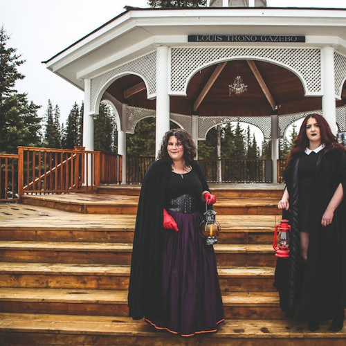 Two women in dark, vintage attire holding lanterns on wooden steps in front of a white gazebo labeled "Louis Trono Gazebo."