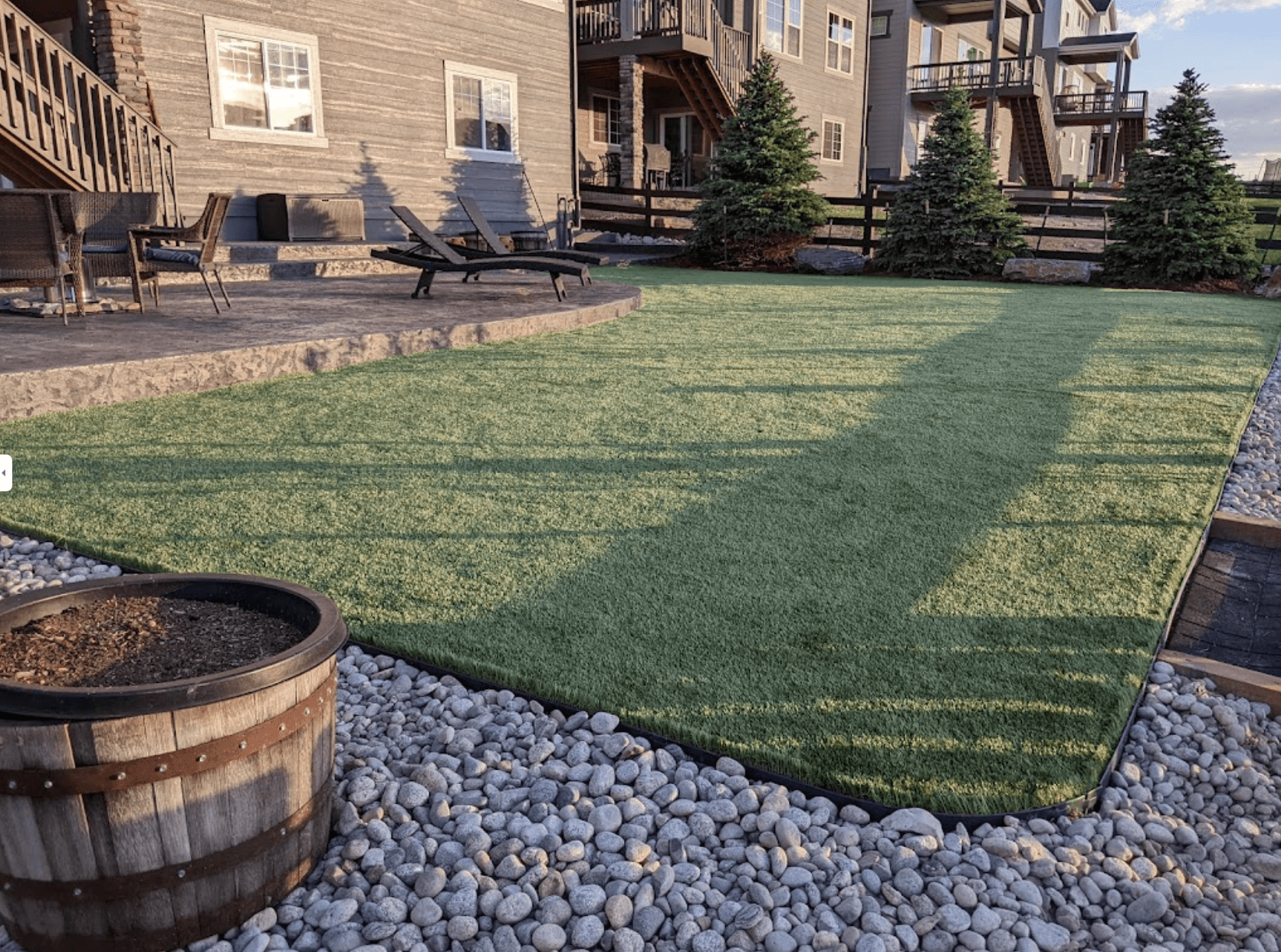 artificial grass installation in a front yard with pebble stones and a wooden barrel for a seating area and firepit