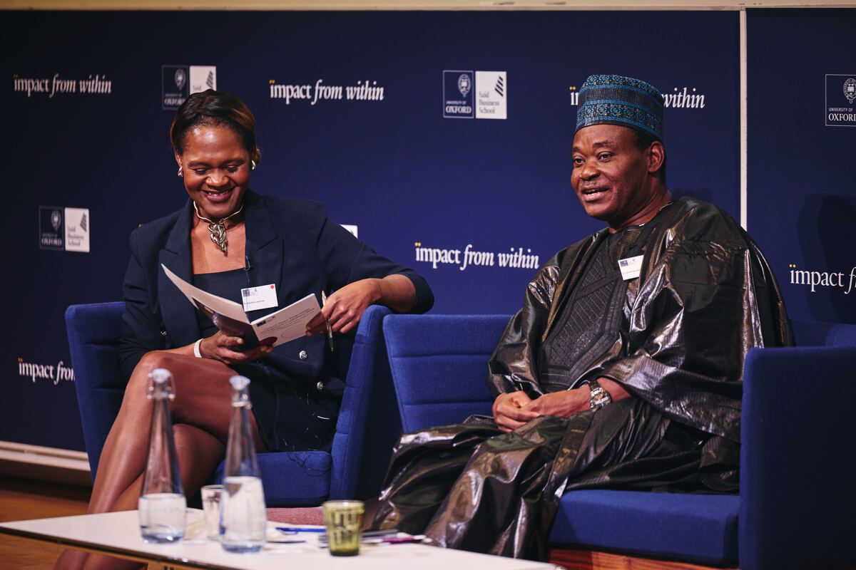 Two panellists in conversation during a seated discussion at the Oxford Major Programme Alliance conference, with an 'Impact from Within' backdrop