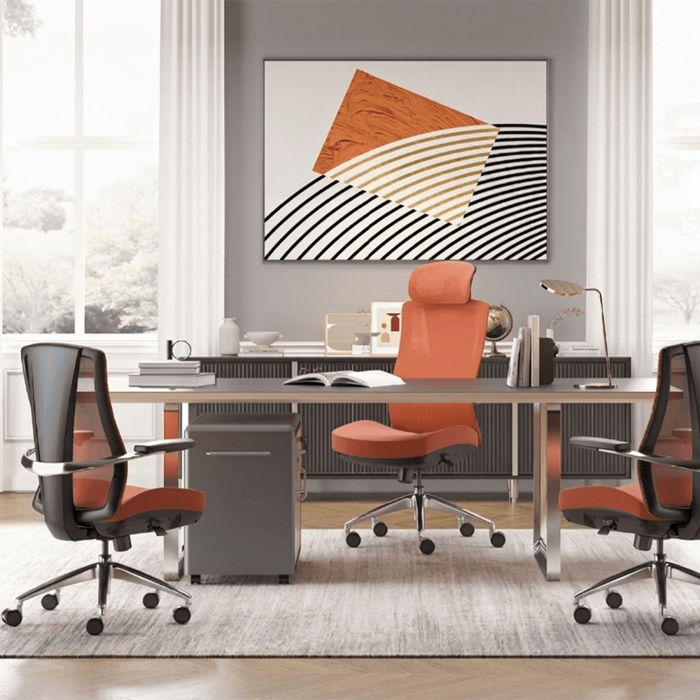 A modern, brightly lit executive office interior. In the center, a large minimalist desk with chrome legs holds an open book and several small office accessories. A high-back ergonomic chair in vibrant orange serves as the main desk chair, flanked by two matching guest chairs in a black and orange mesh design.  Behind the desk is a dark grey credenza topped with books and decor, positioned under a large, abstract piece of wall art featuring geometric shapes in orange, white, and black. To the left, a tall, slender floor lamp with a spherical glass shade stands near a window with white curtains, while a potted green plant sits to the right. The room features light wood herringbone floors and a soft, grey textured rug.