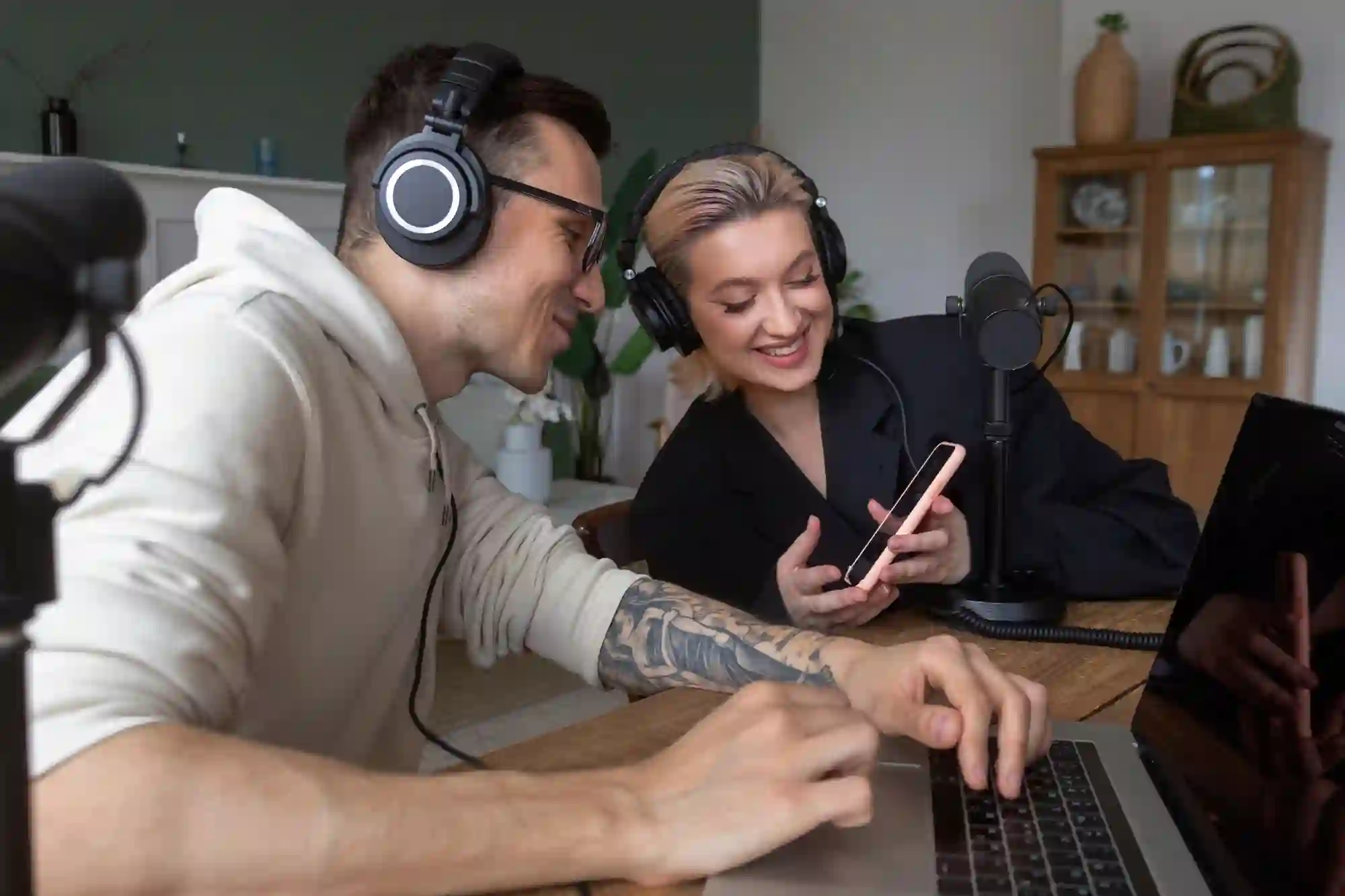 A tattooed man and a woman with a phone are seated at a table, both wearing headphones and smiling while working on a laptop with podcast microphones set up.