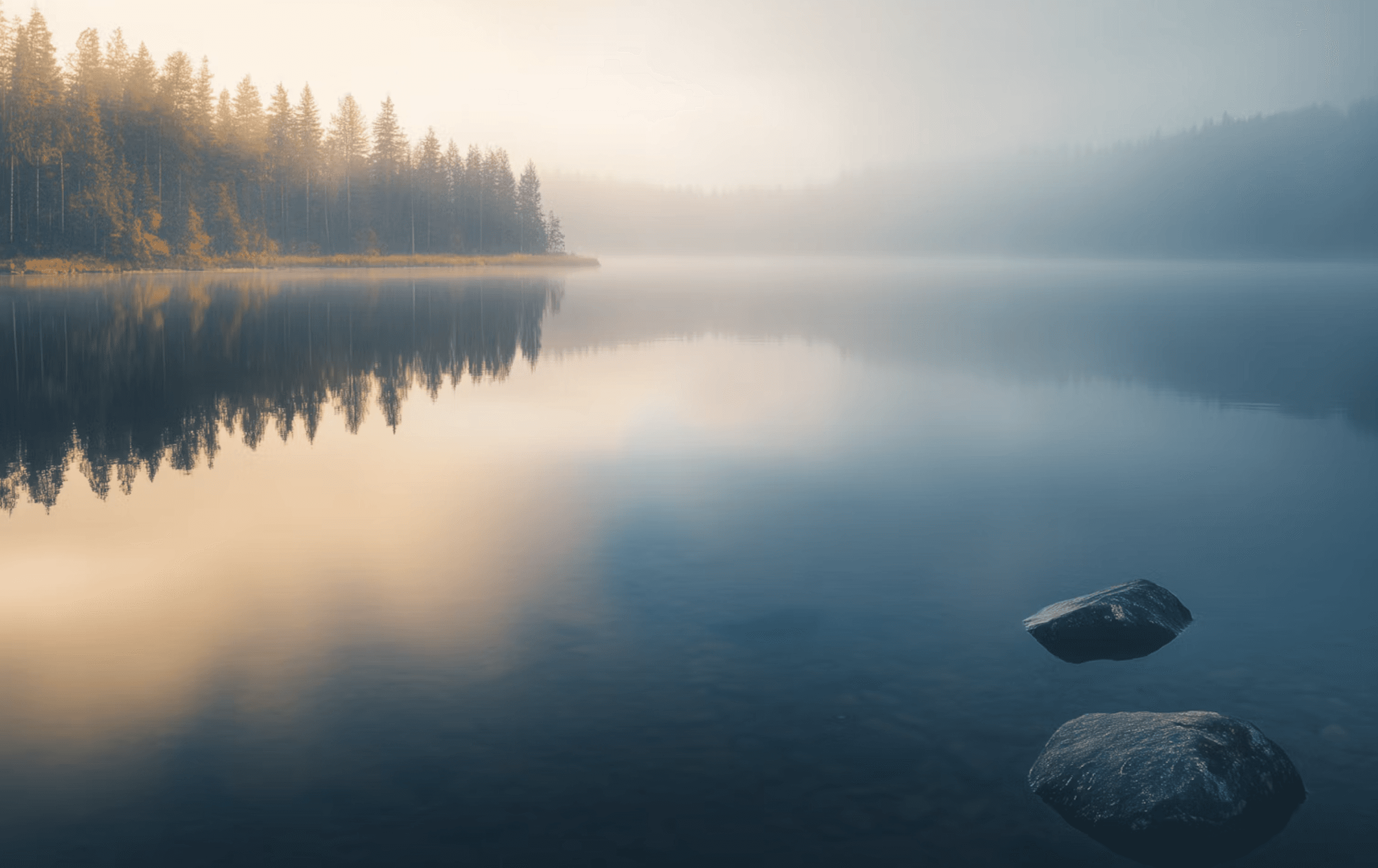 Peaceful lake at sunrise with misty reflections of trees, golden light, and smooth rocks in the foreground. A serene, dreamy landscape