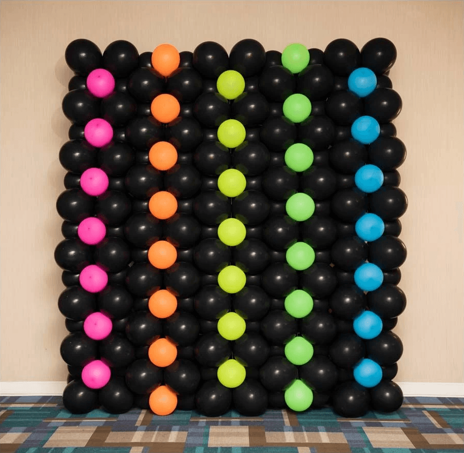 A vibrant and colorful balloon wall art installation features black balloons arranged in a grid pattern, accentuated with vertical rows of bright neon balloons in pink, orange, green, and yellow, creating a striking and festive visual effect.
