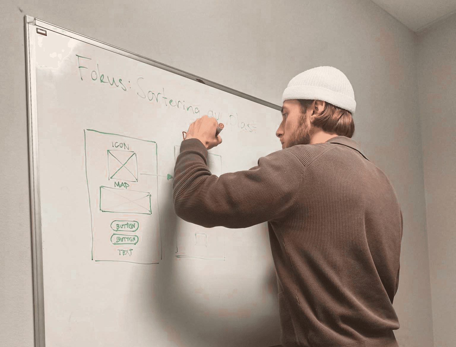 Lukas wearing a white beanie and brown sweater, is writing on a whiteboard with a red marker. The whiteboard contains a rough wireframe sketch for a digital interface, including labeled elements such as icon, map and button