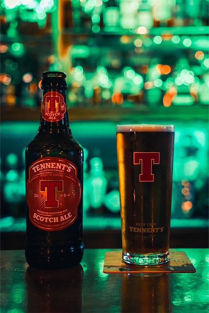A bottle of beer next to a pint glass on a bar, with green lighting and blurred shelves of bottles in the background.