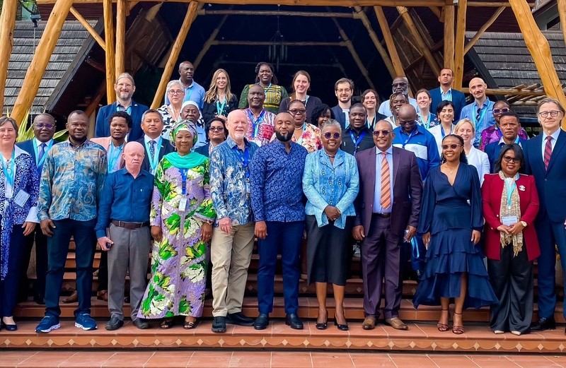 A wide group photograph of international delegates and African leaders standing together on tiled outdoor steps at the 11th Our Ocean Conference in Mombasa, Kenya. The large group is dressed in a mix of formal business suits, smart-casual attire, and vibrant traditional African garments. They are posed in front of a striking, prominent entrance featuring an intricate, vaulted bamboo and timber roof structure.