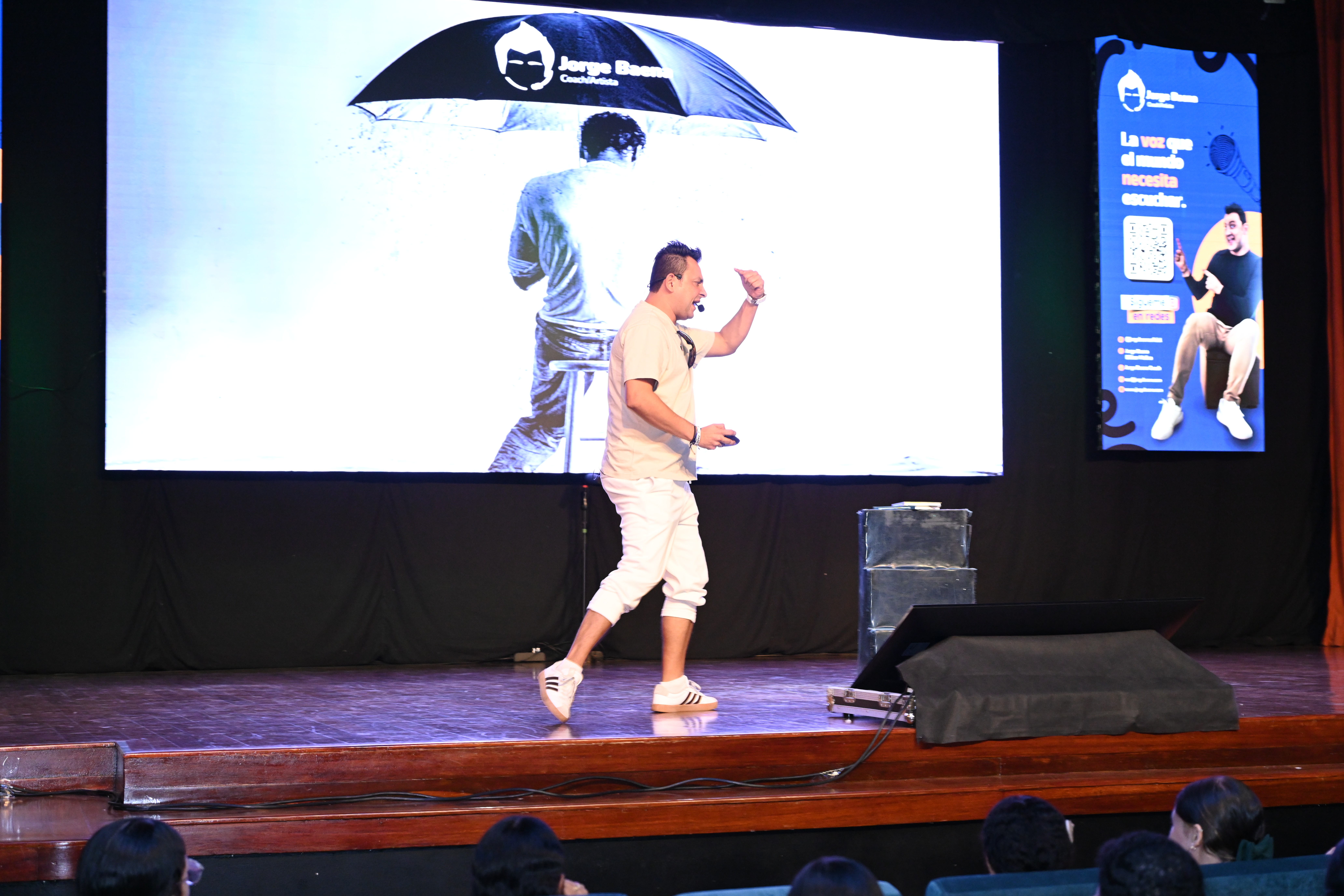 Jorge Baena en el escenario durante una conferencia, caminando frente a una gran pantalla que muestra la imagen de una persona bajo un paraguas con el logo de su marca, mientras se dirige al público con energía.