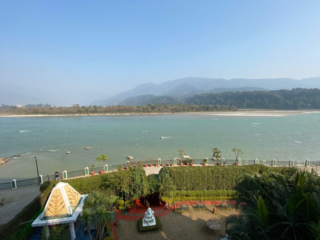 The river Ganga flowing alongside the mountains in the background and a small temple in the foreground.
