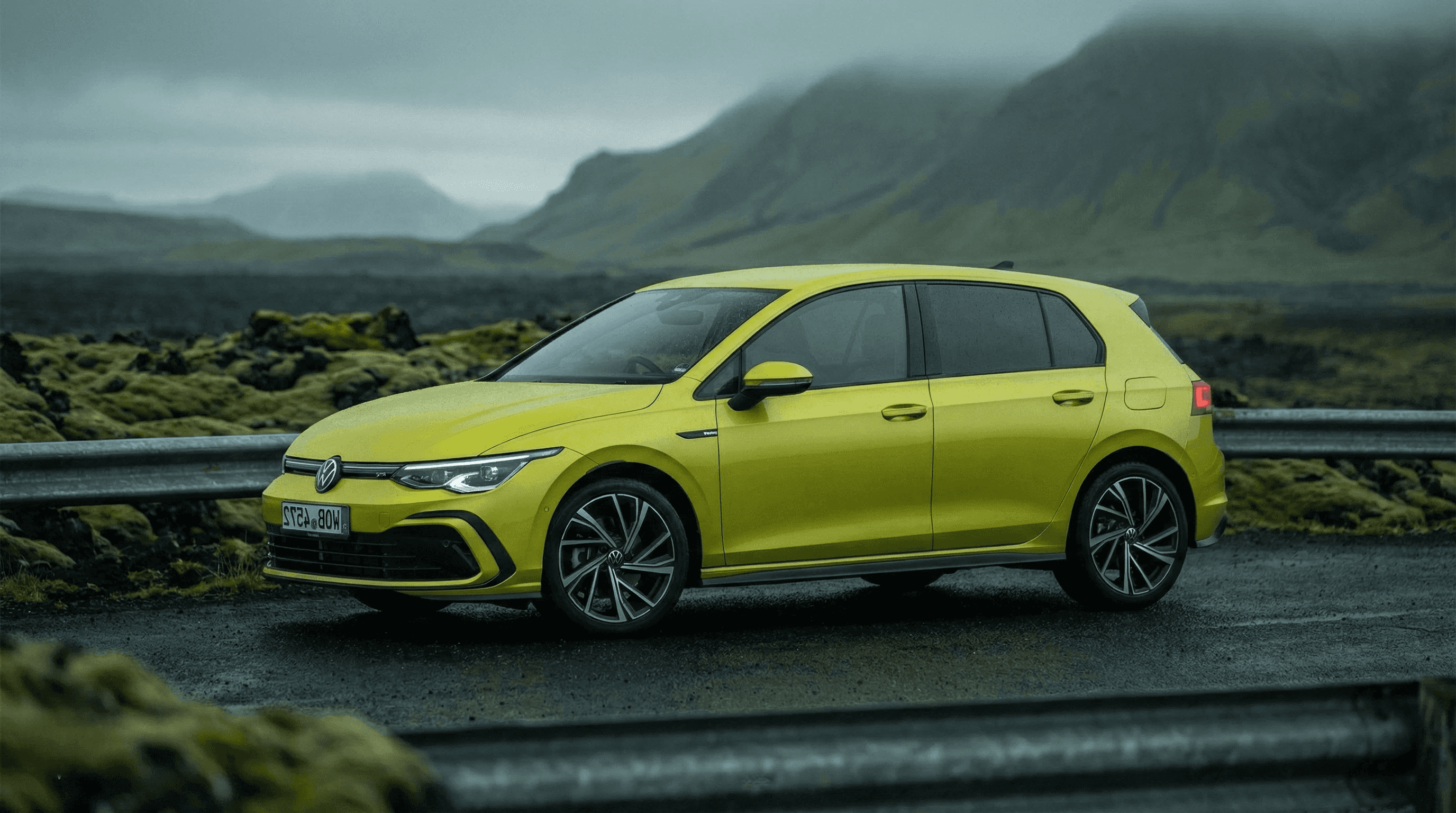 A bright yellow VW Golf parked on a road with green mossy hills and a cloudy sky in the background.