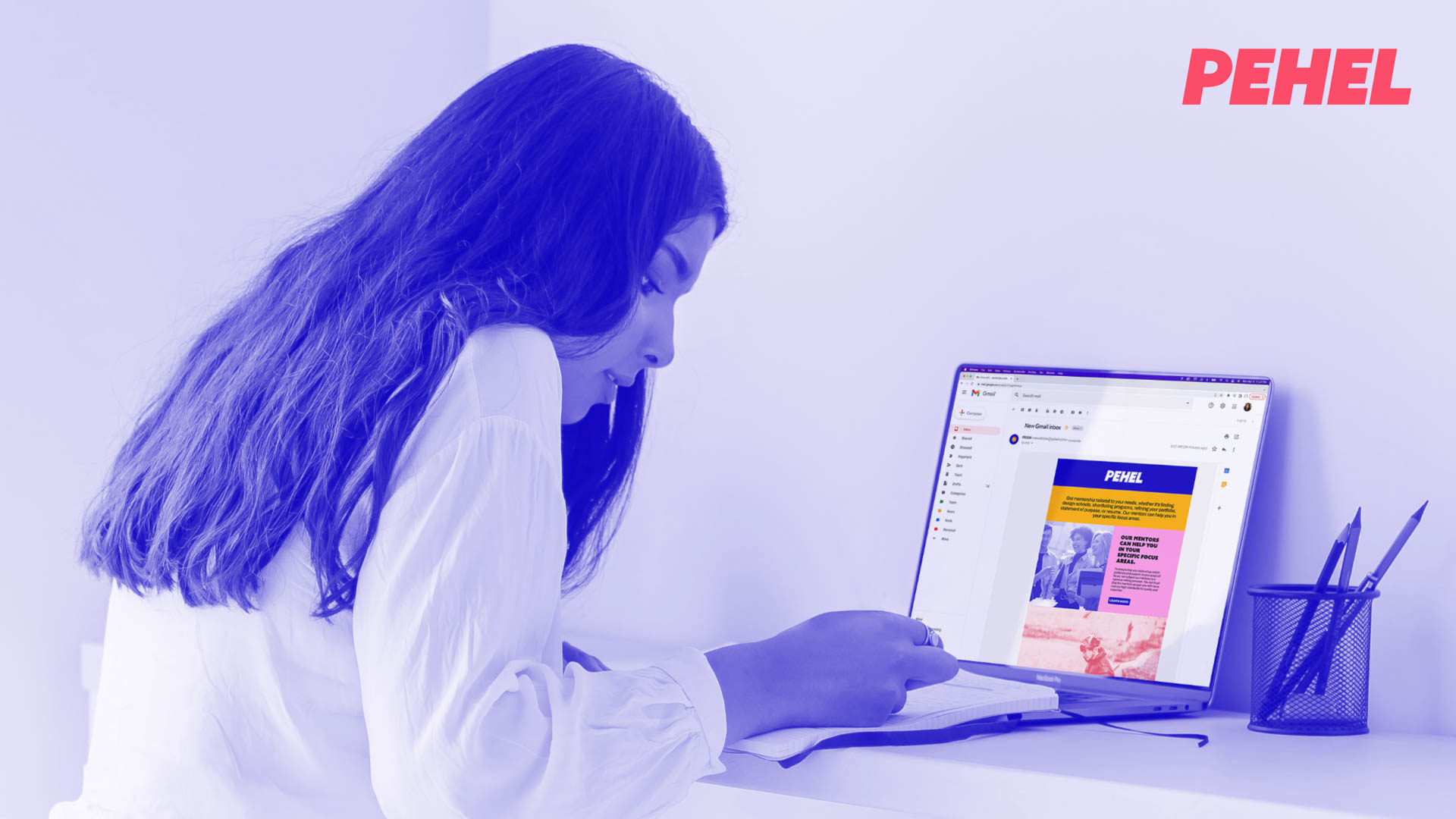 A woman with long dark hair, wearing a white long-sleeved shirt, is seated at a white desk and looking at a laptop screen displaying the PEHEL website. The laptop is open and positioned in front of her. The background is a light purple or lavender color. To the right of the laptop, a black mesh pencil holder with pens is visible. The PEHEL logo is prominently displayed in the top right corner of the image.