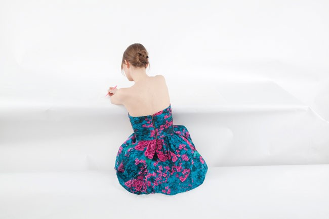 A woman with brown hair tied in a bun sits on a white surface, facing away from the camera. She wears a strapless dress with a teal and pink floral pattern and a pink bow at the waist. Her right hand holds a pink object near her face. The background is entirely white.