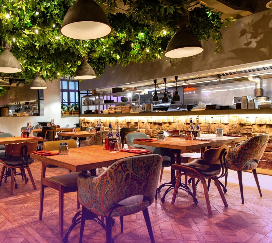 Dining area of a modern restaurant with wooden tables, chairs, and greenery hanging from the ceiling.