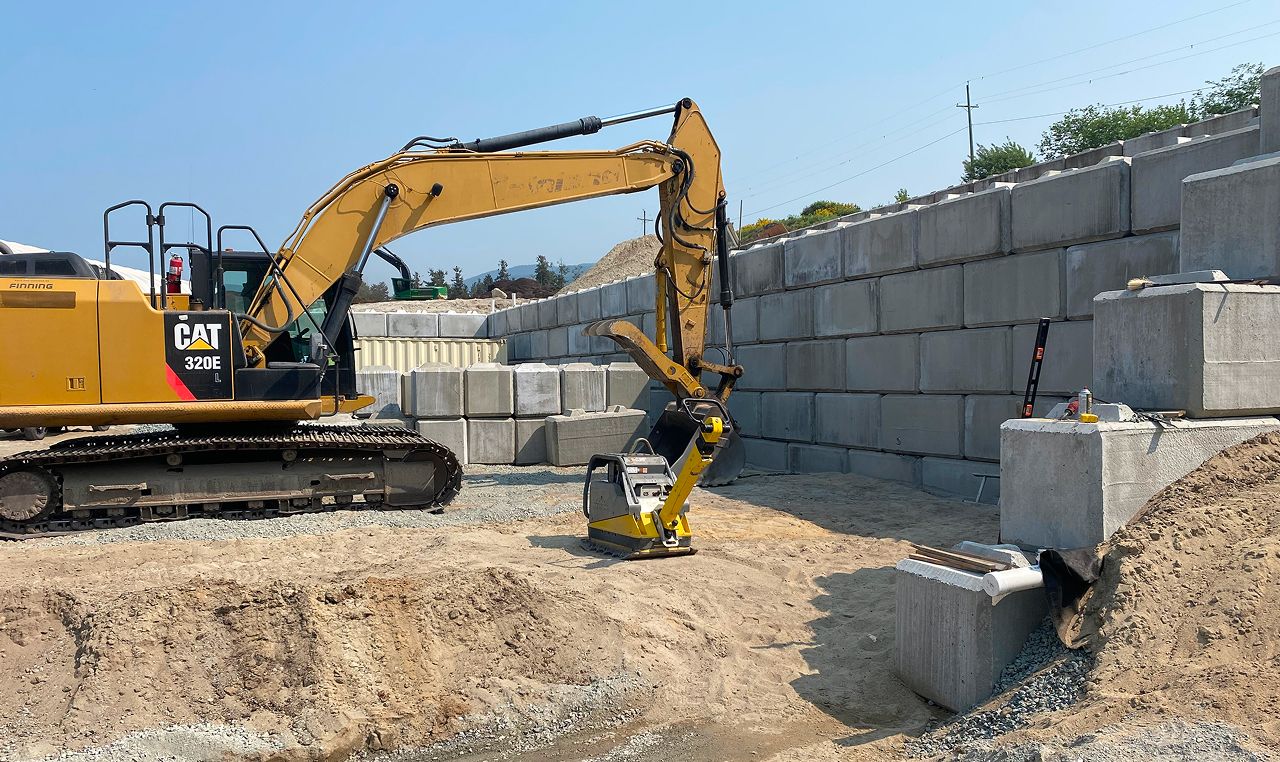 Excavator compacting backfill during lock block retaining wall construction at Salish Soils facility