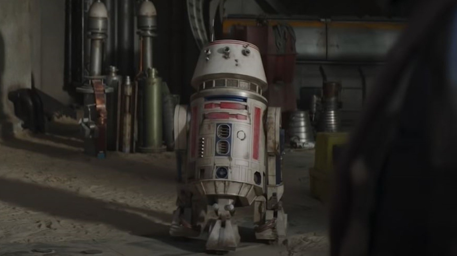 A small, round robot with a dome-shaped head and various mechanical details, standing in a dimly lit setting.