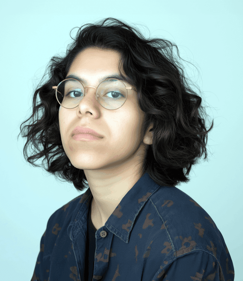 A woman with glasses, dressed in a shirt, smiling softly while standing against a plain background.