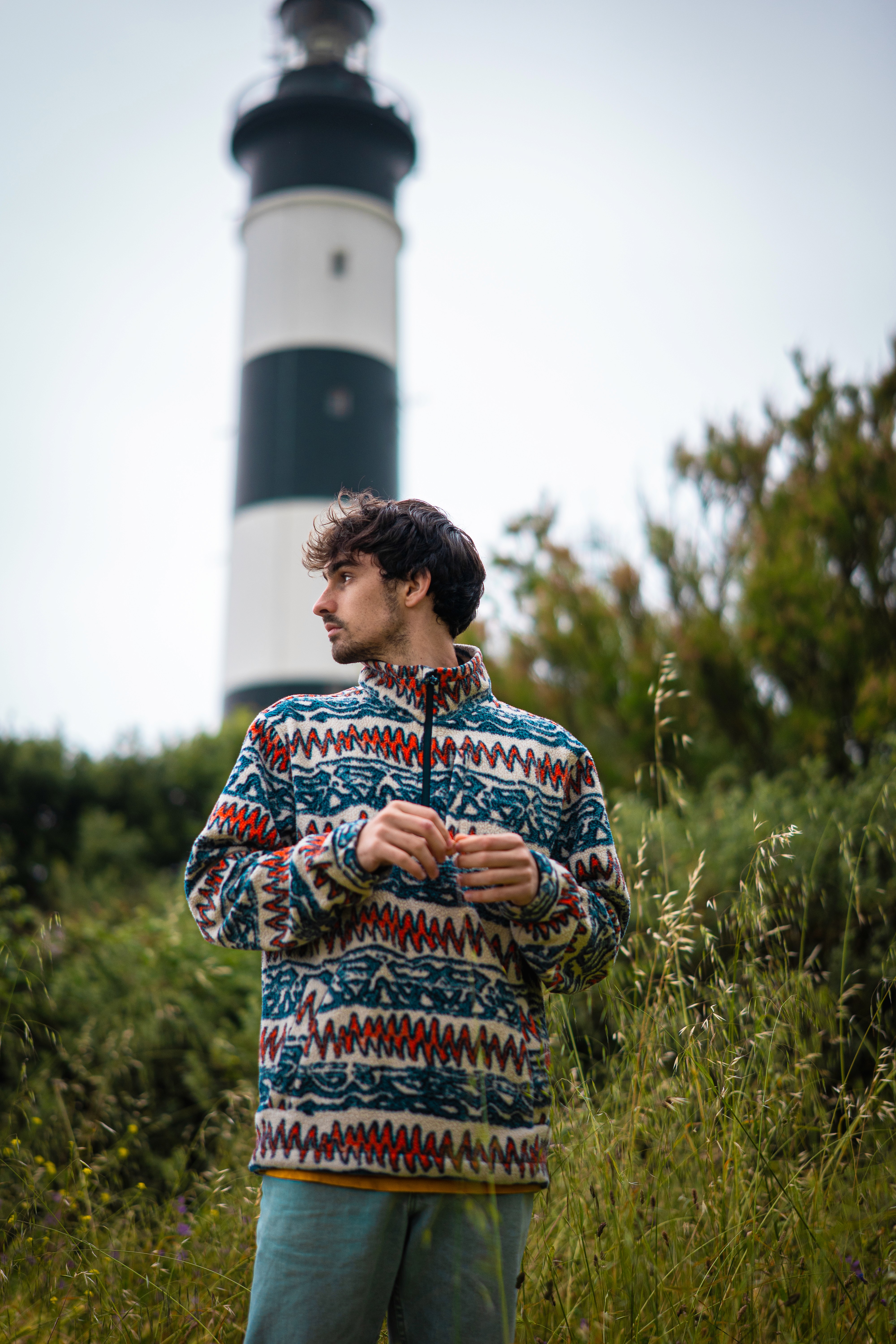A side-profile of a young person with dark, wavy hair, wearing silver sports sunglasses and a black sweater, sitting in front of a blurred teal couch.