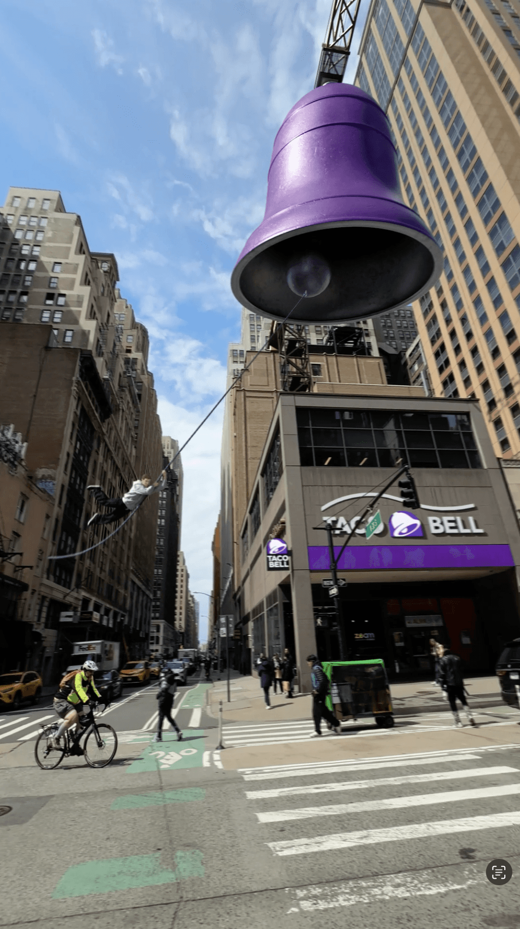 A giant purple bell hangs over a city street while a person swings from its rope near a Taco Bell