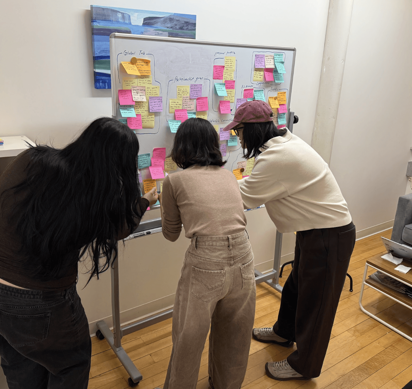 Three team members standing at a whiteboard covered in colorful sticky notes, mid-affinity-mapping. Notes are clustered around hand-written category labels.