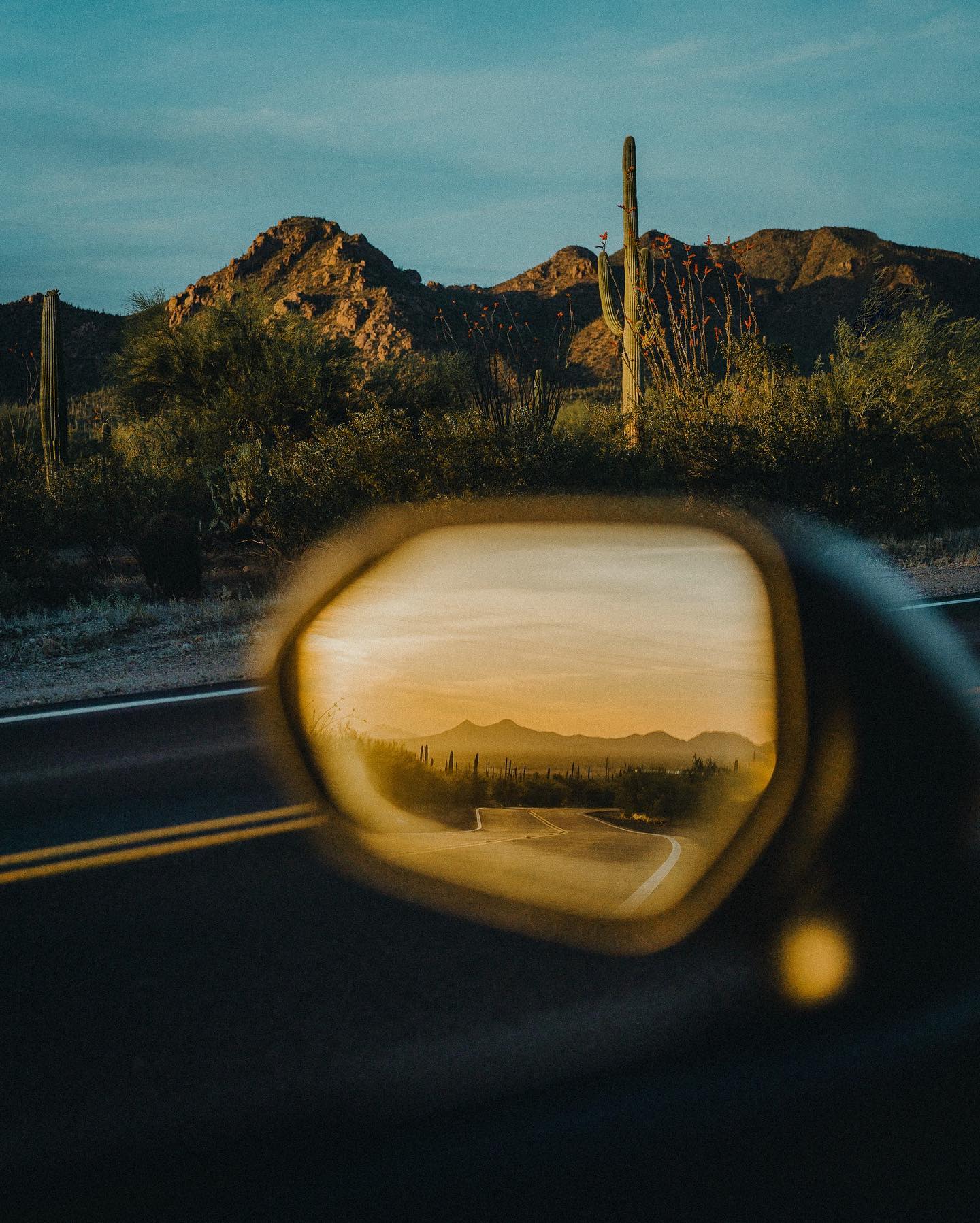 Desert road reflected in the side mirror of a car with a Saguaro cactus and mountain in the background.