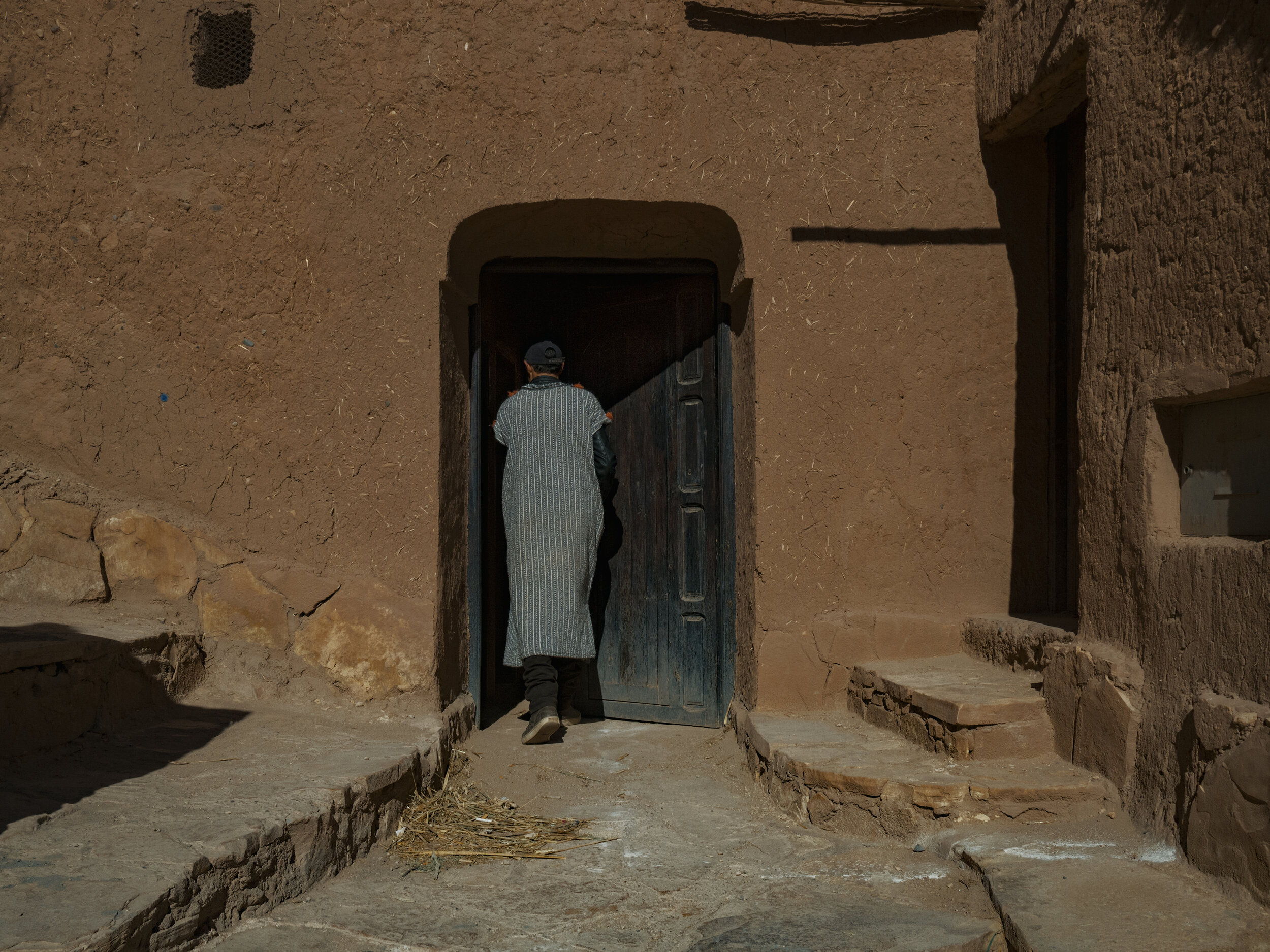 Back view of a person entering an earthen building in a rustic Moroccan village, wearing a striped robe and cap.